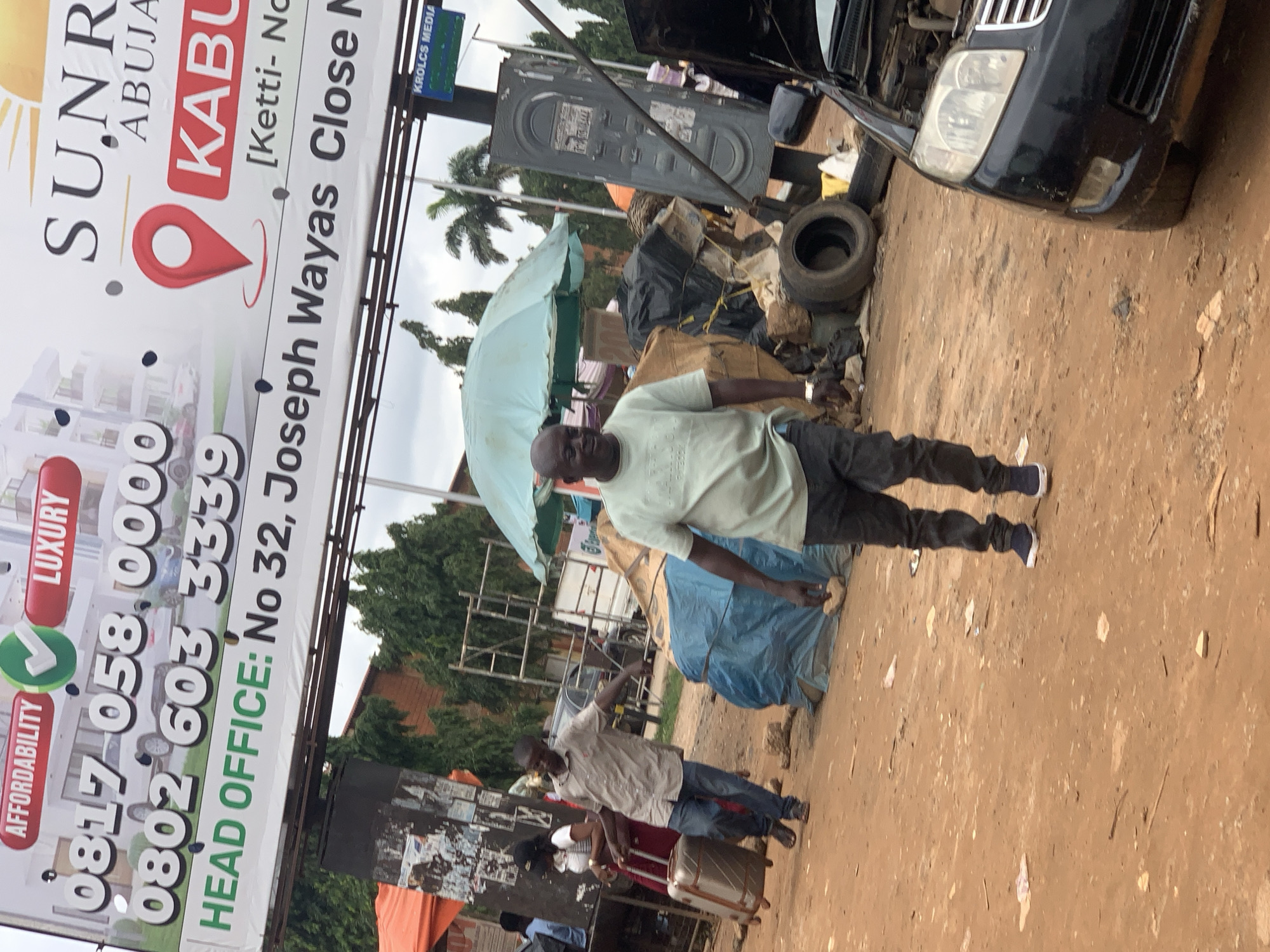 The image shows a street scene in Abuja, Nigeria. In the background, a large billboard advertises "SUNRISE", "ABUJA", and "KABU". It also displays phone numbers "0817 058 0000" and "0802 603 3339", and the text "HEAD OFFICE: No 32, Joseph Wayas Close". In the foreground, a man in a light-colored shirt and dark pants is walking and pulling a suitcase. To his right, another man in a light green shirt and dark pants is walking towards the camera. There is also a partially visible black car with its hood open, and various items scattered on the ground including tires and debris. The ground is dirt and appears to be unpaved. In the middle ground, there is a blue tarp covering some items, and a light blue umbrella.