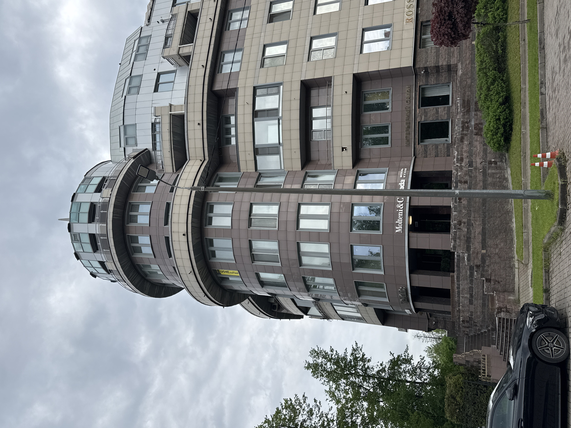 The image depicts a contemporary building located in Saint Petersburg, Russia. The building has a unique, curved design with a mix of brown and silver-colored paneling. It appears to be a commercial building with signage indicating businesses such as "Molteni&C | Dada," and "ROSSI." A black car is parked near the building's entrance, and there are traffic cones placed along the curb. The sky is overcast, and trees are visible in the foreground. The overall environment is urban with well-maintained landscaping.