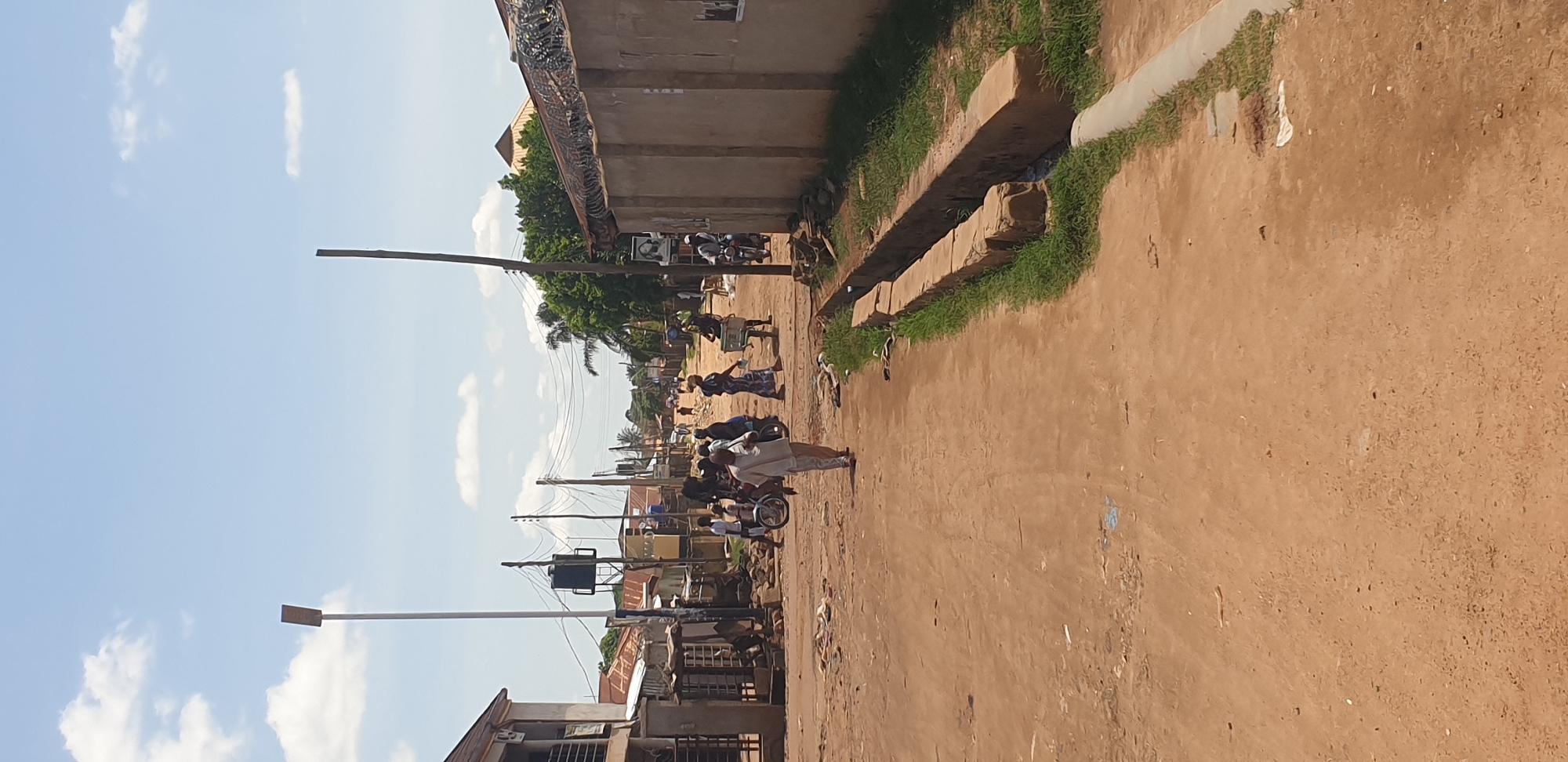 This image depicts an unpaved, reddish-brown dirt road in Sabon Tasha, Nigeria, under a bright, clear sky with scattered white cumulus clouds. The scene is illuminated by strong sunlight, casting visible shadows.
On the left side of the road, a two-story building with a brown roof is partially visible, featuring a window and an obscured entrance. Further down, multiple utility poles support a dense network of overhead wires. A black elevated water tank on a metal frame stands amidst other smaller structures, some with corrugated metal roofs. A tall, light-colored pole with a rectangular, flat light fixture or solar panel extends into the upper left quadrant of the sky.
On the right side of the road, an open concrete drainage ditch, partially filled with green grass, runs parallel to the road. Beyond the ditch, a high, light-colored concrete wall topped with coiled razor wire extends into the background. A dark-framed portrait, appearing to depict a human face, is affixed to this wall. A tall, dark utility pole stands vertically on this side. Green foliage from trees and bushes is visible behind the wall and utility pole.
Several individuals populate the road. In the foreground-midground, an adult male wearing a light-colored long-sleeved shirt and trousers walks towards the viewer, slightly right of the road's center. Further down the street, multiple other figures are visible. Some individuals appear to be carrying objects on their heads or in their hands. At least two motorcycles or mopeds are present on the road, each with a rider. The road recedes into the background, where more buildings and trees line the street under the distant sky. The overall environment suggests a busy, semi-urban thoroughfare.