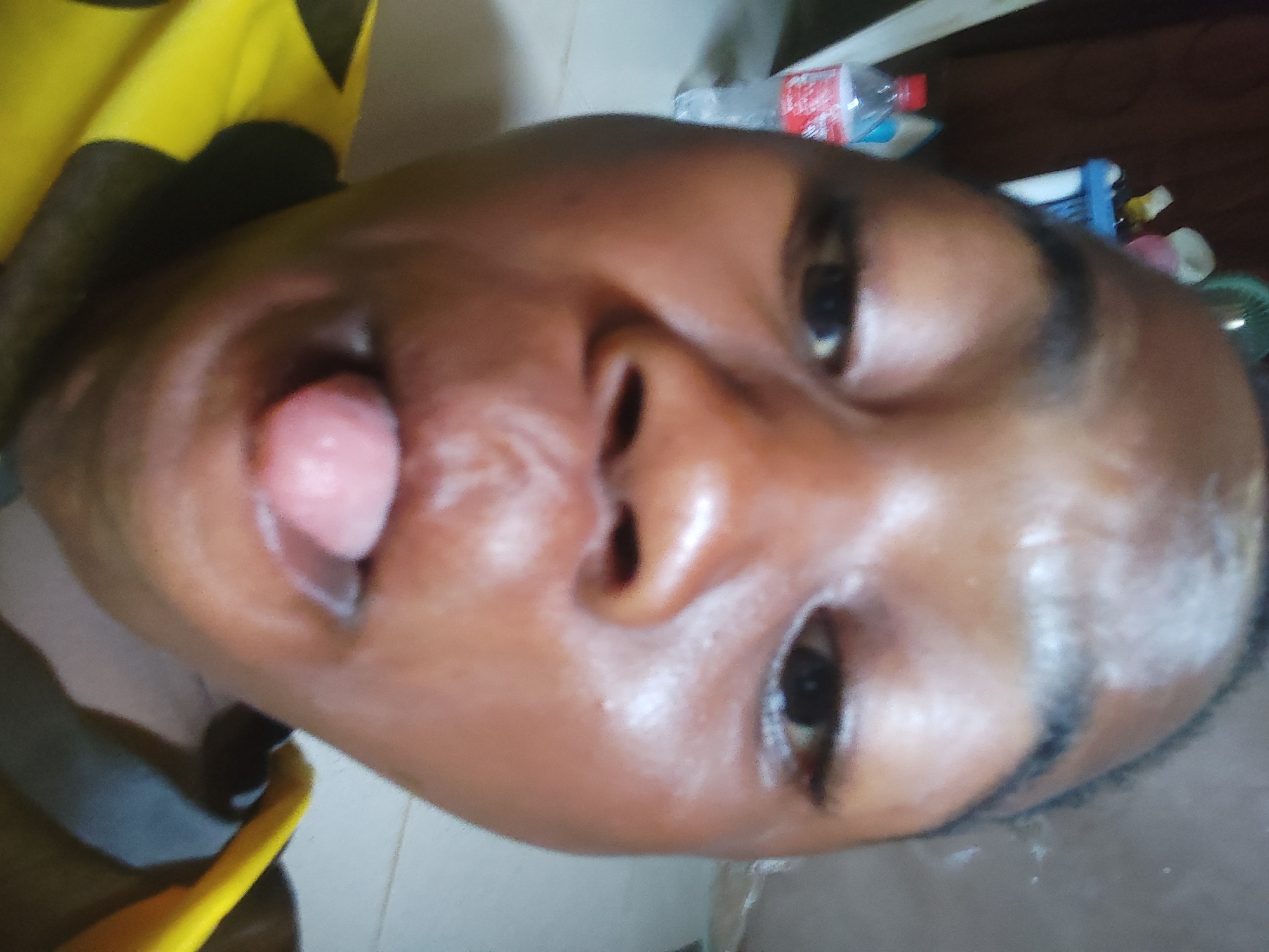 A close-up photograph shows a person with dark skin and dark eyes sticking their tongue out. The background is blurry, but various objects including a water bottle are visible. There is no concrete evidence to confirm the location as Ikorodu, Nigeria.