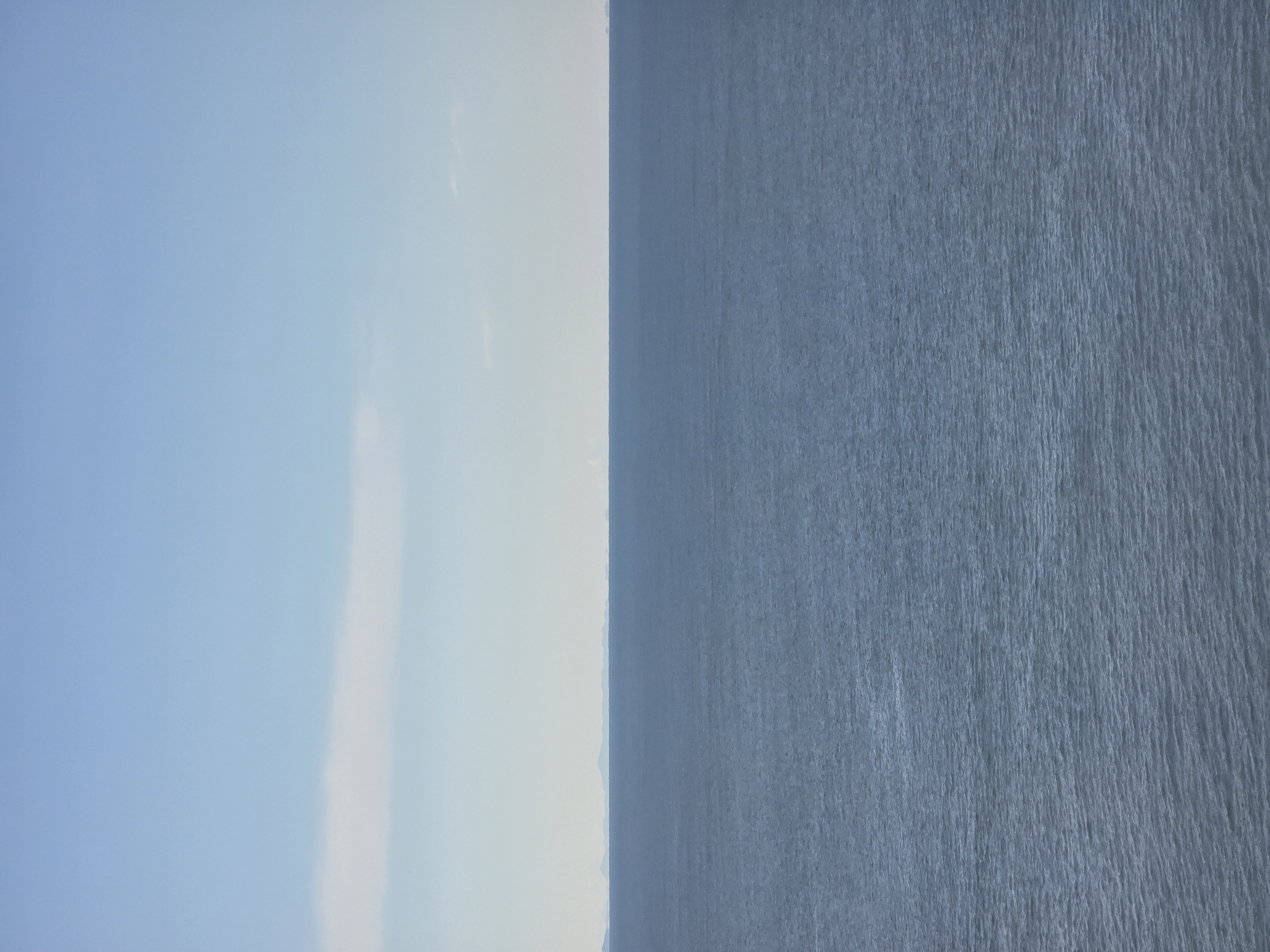 The image shows a calm sea meeting the sky, possibly captured in Ulcinj, Montenegro, due to its geographical location. The left side of the image features a clear, light blue sky, while the right side depicts the water with slight ripples on the surface. The horizon appears as a line that separates the two, and there are no visible people or objects in the water or sky.