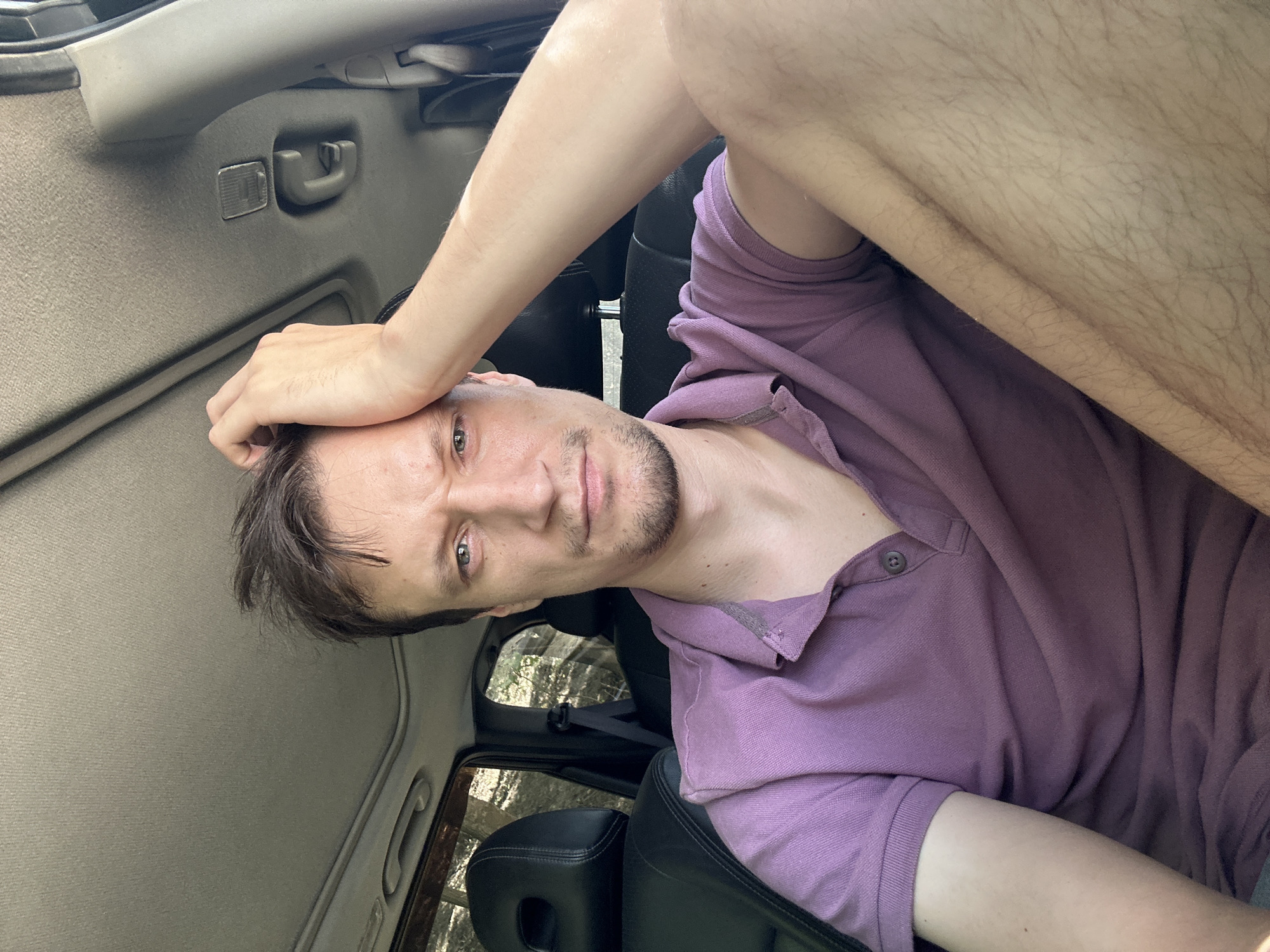The image shows a young man with short brown hair, fair skin, and a light beard, sitting in the driver's seat of a car. He is wearing a purple polo shirt. His right hand is resting on his forehead, and he is looking at the camera with a concerned expression. The interior of the car is visible, including the sunroof, dashboard, and leather seats. The background appears to be a wooded area, suggesting the car is parked or driving in a rural setting. The lighting indicates it is daytime. The location is not discernible from the image itself.