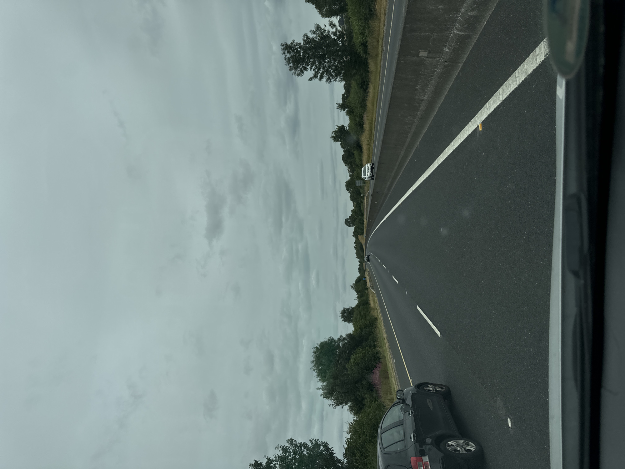 An overcast sky looms over a multi-lane highway in Ballyduff South, Ireland. The road is paved with dark asphalt, marked by white lines. A black hatchback is in the left lane, closest to the viewer. In the right lane, a white SUV is visible further down the road. The sides of the highway are lined with green vegetation and trees. A concrete barrier separates the lanes of traffic. The scene suggests a typical day of travel on a major road in the Irish countryside.