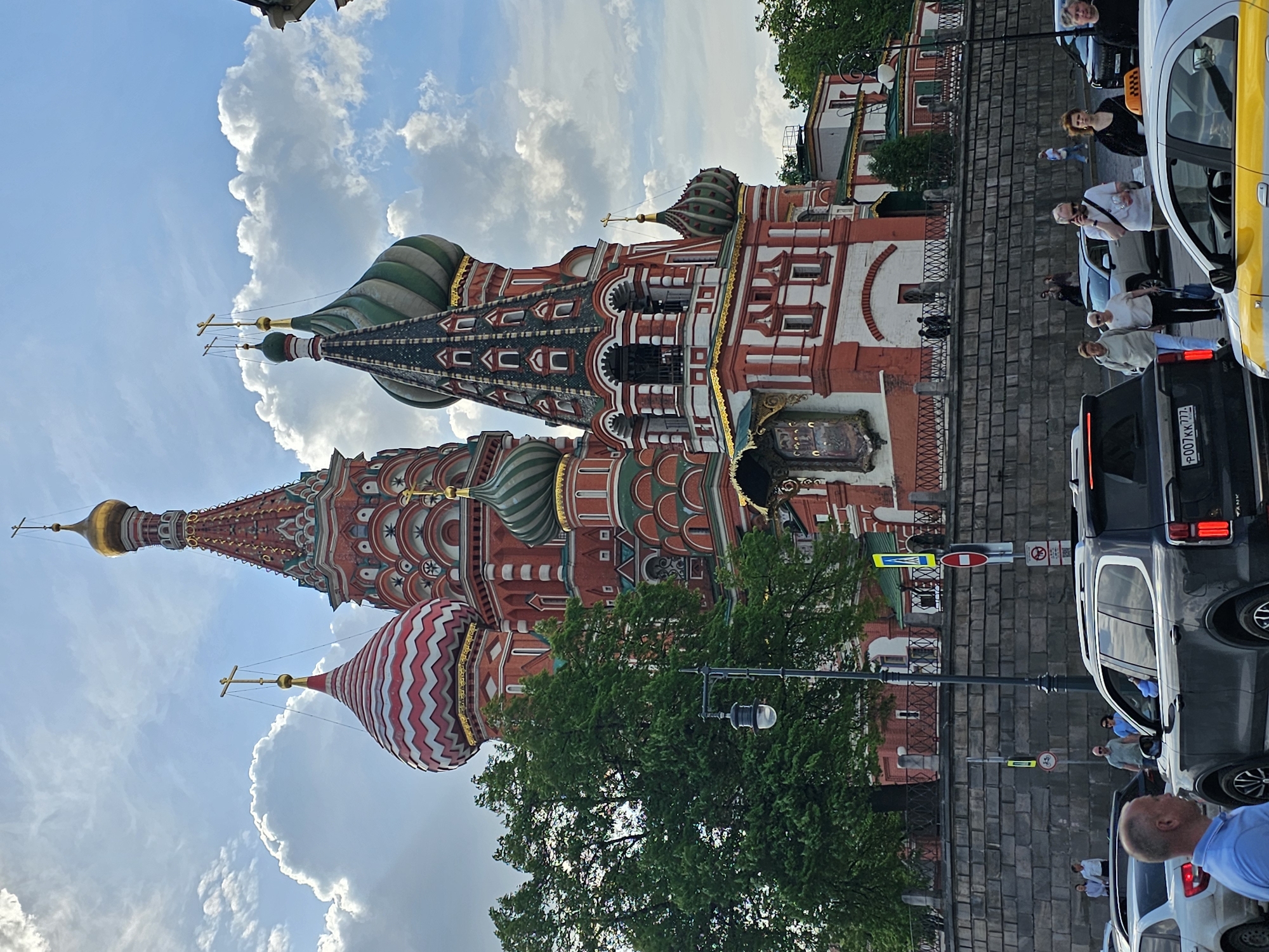 The image shows St. Basil's Cathedral in Moscow, Russia, a multi-domed Orthodox church with vibrant colors and patterns. The architecture features a variety of domes topped with crosses. Below the Cathedral, there is a street with people, cars, and traffic signs. The sky is visible with clouds.