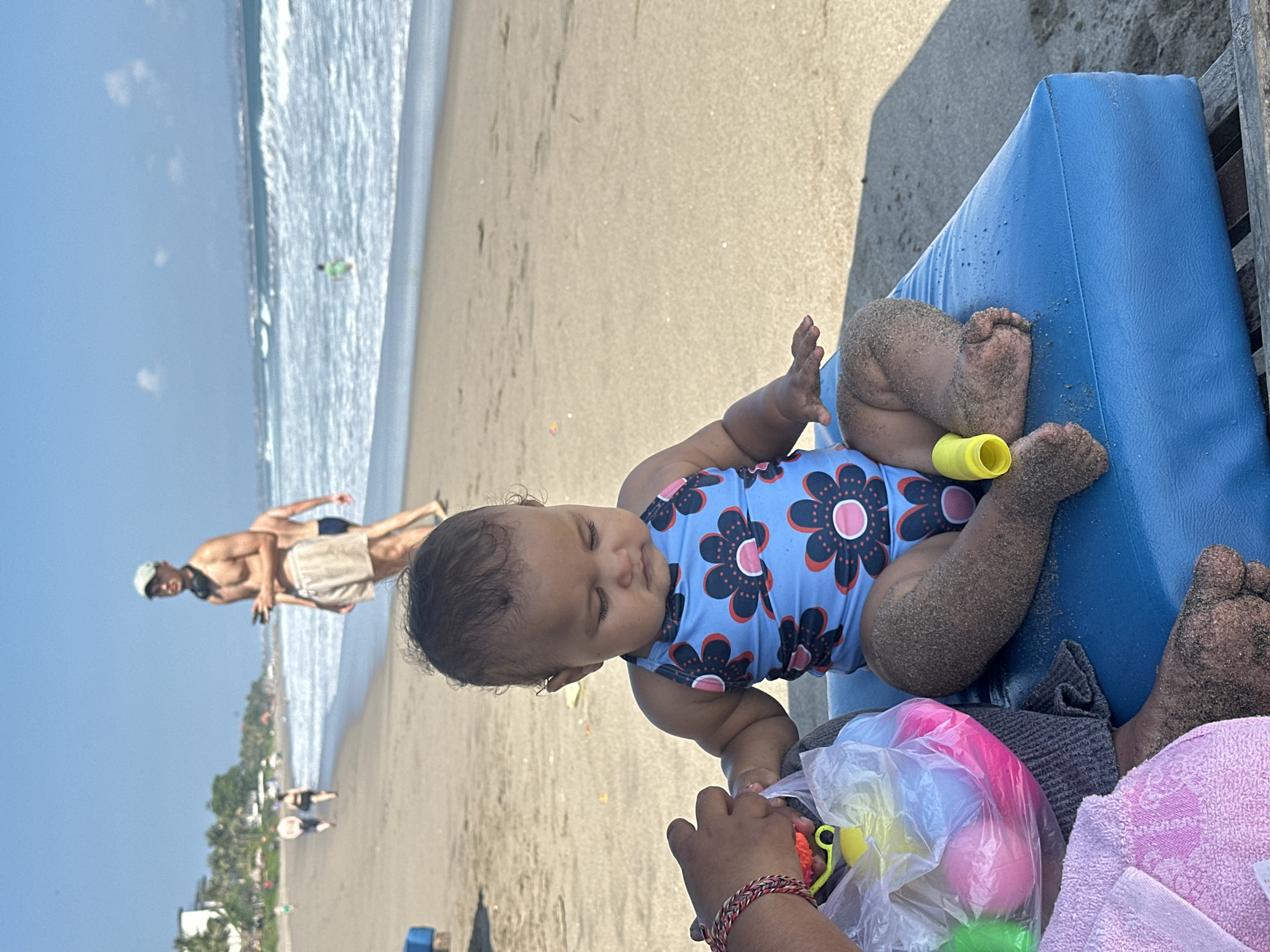 An outdoor, medium-shot, eye-level, vertical photograph captures a baby sitting on a blue beach chair on a sandy beach in North Kuta, Indonesia. The baby is wearing a blue swimsuit with a floral pattern in pink and black. The baby's legs and feet are covered in sand. To the left of the baby, a person's hand is holding a clear plastic bag filled with pink and yellow toys. The background shows the ocean with waves, a sandy shoreline, and a few people in the distance walking and wading in the water. The sky is clear and blue. A man in the mid-ground, facing left, is wearing light-colored shorts and a cap, with headphones around his neck. The overall lighting suggests a sunny day.
