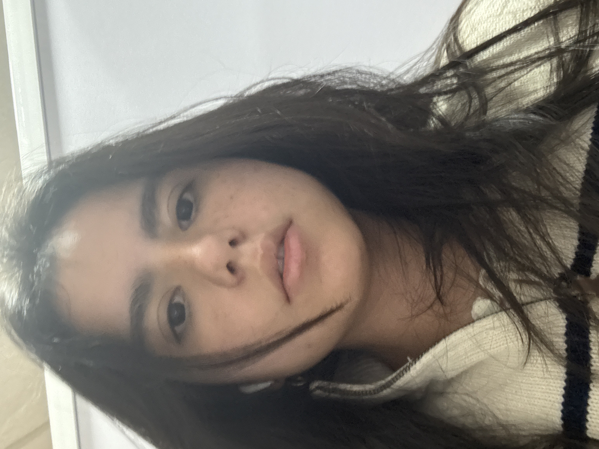 A close-up, slightly angled self-portrait of a young woman with dark hair. She has fair skin with some freckles. She is wearing a cream-colored sweater with dark blue stripes. A white object, possibly an earbud, is visible near her ear. The background appears to be a white wall or surface. The photograph was taken indoors. There is no location information for Metepec, Mexico included.