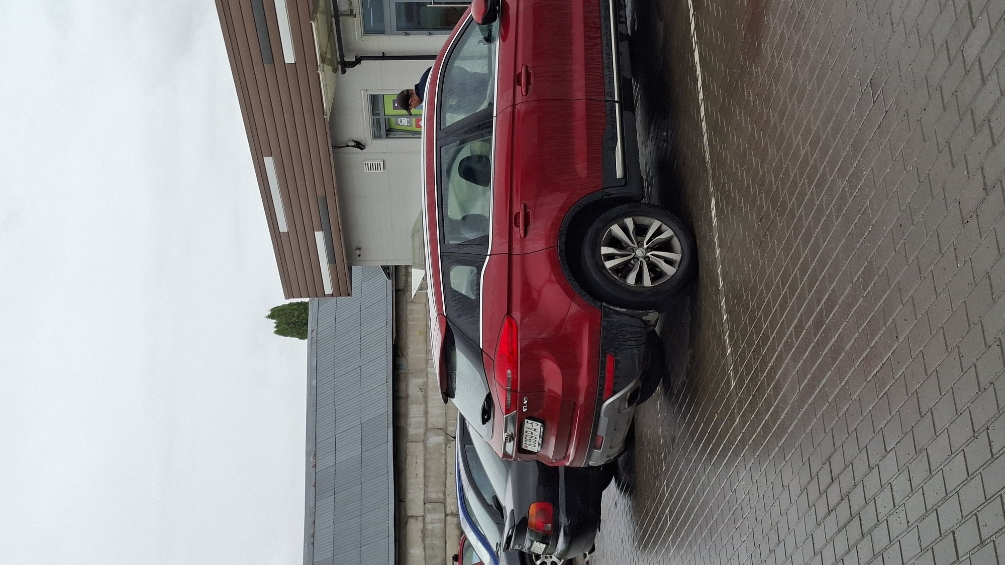 In the city of Smolevichi, Belarus, a parking lot is depicted on a cloudy day. The ground is paved with dark, wet interlocking bricks. A red SUV, identified by its license plate as 76359 PA-5, is parked in the foreground. To the left of the red SUV, a black sedan is partially visible. In the background, a building with a brown and white striped facade is seen, and behind that, a grey corrugated metal roof structure. A single tree stands to the left of the building. A person wearing a dark jacket and a cap is standing near the entrance of the building.