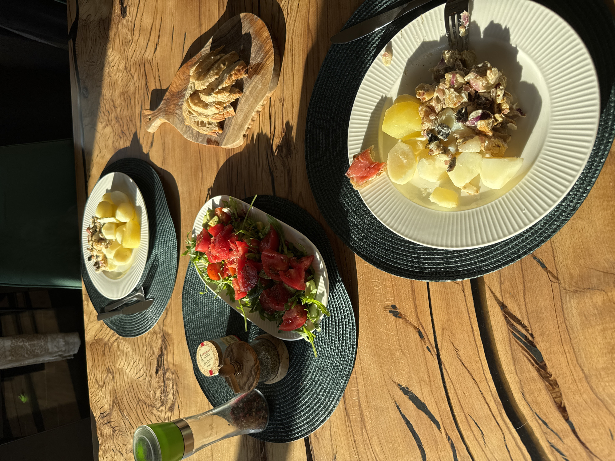 The image shows a table with a meal laid out on it. The meal consists of sliced bread on a wooden board, two plates of potatoes with a topping, and a plate of tomatoes and greens. There are also condiment bottles and a pepper mill on the table. The table is made of wood and has a visible grain pattern. The lighting suggests it is daytime, and the scene is indoors. There are no people visible in the image. The location information is not discernible from the image.