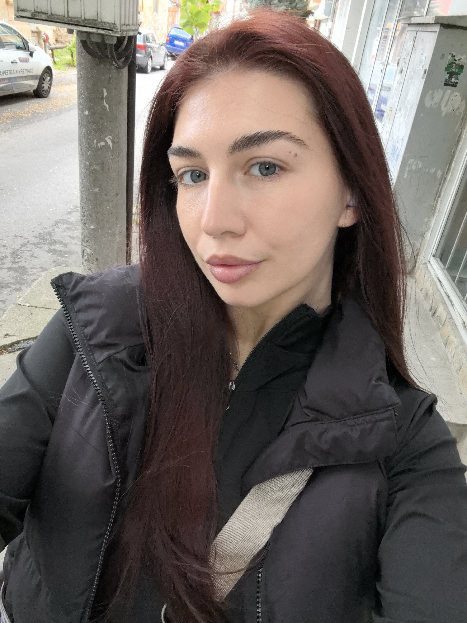 A young woman with long, dark reddish-brown hair is taking a selfie. She has fair skin and blue eyes. She is wearing a black puffer vest over a black shirt, and a light-colored strap (possibly for a bag or purse) is visible across her chest. She appears to be wearing wireless earbuds. The location is suggested to be Stara Zagora, Bulgaria.
Behind her is a sidewalk with a grey concrete utility pole to her left. Parked cars are visible in the background on the street. There is a building to her right with a partially visible window display. The weather appears overcast.
