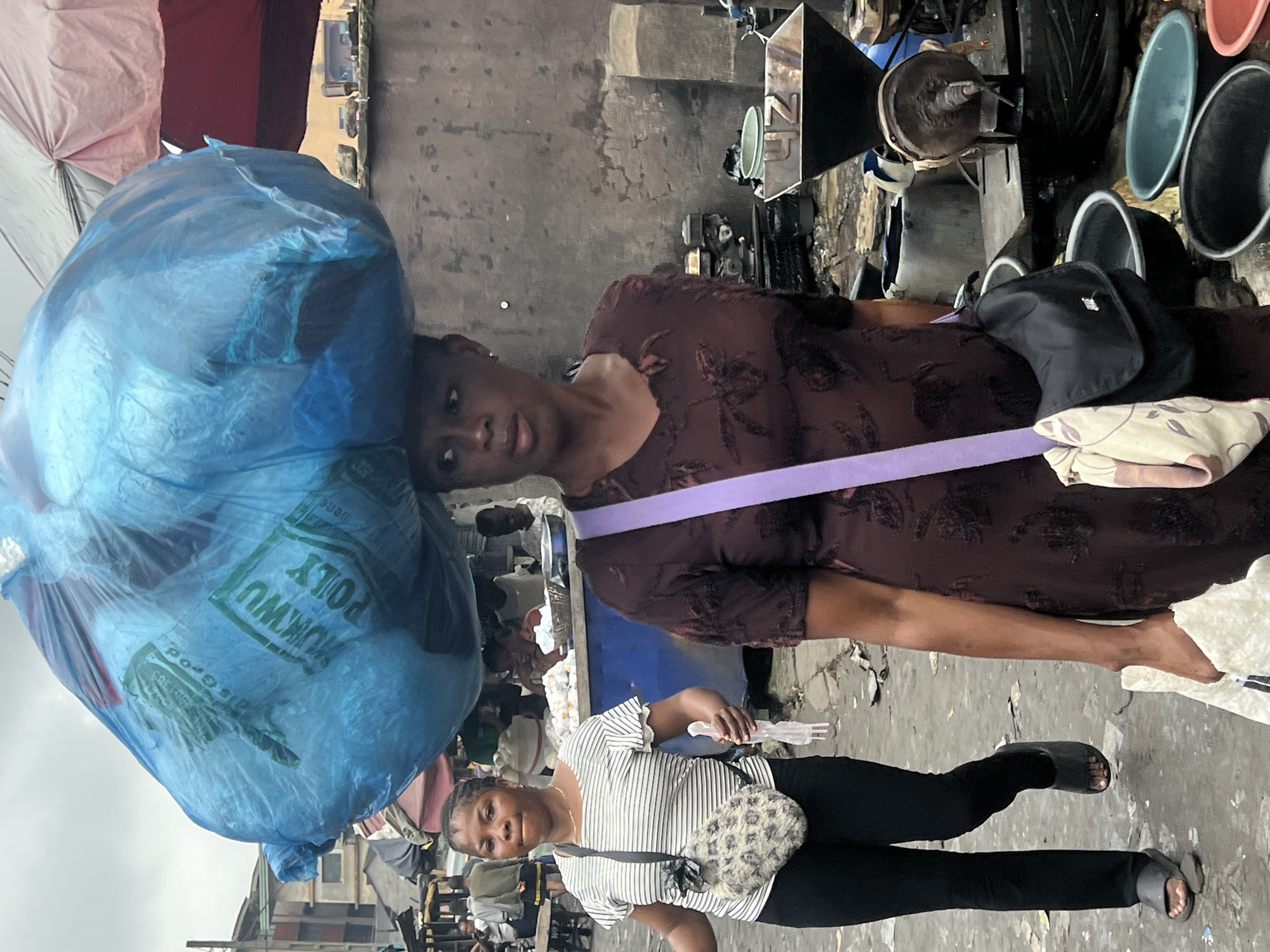 In the city of Nkpor, Nigeria, a woman walks under a cloudy sky, carrying a large, blue plastic bag filled with goods on her head. She is wearing a dark, patterned dress and a purple strap from a bag crosses her chest. In the foreground, another woman walks to the left, wearing a striped t-shirt and black pants. She is carrying a leopard-print bag across her body and holds a white plastic object in her right hand. The background suggests a bustling market scene with stalls, umbrellas, and various objects, including a metal weighing scale with the numbers "4Z" visible on it. The ground is dirty and littered with debris.