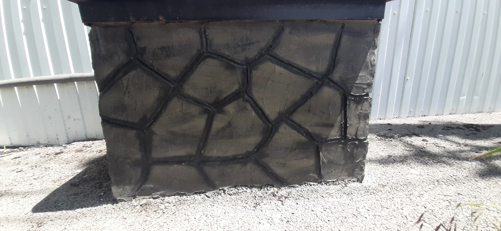The image shows a close-up, eye-level view of a dark, textured stone-like structure. The structure appears to be a planter or the base of a column, with a black rim visible at the top. The surface is imprinted with a pattern resembling irregular, polygonal stones, outlined in black. The texture of the stone is rough and uneven, with variations in shade suggesting depth and age.
The structure is set against a background of a white, corrugated metal fence. The fence has vertical seams and appears to be outdoors, as indicated by the gravelly ground beneath the structure and the shadow cast to its left. A thin, dark horizontal pipe or bar runs across the frame in front of the fence, partially obscured by the structure.
The lighting suggests a bright, sunny day, with shadows falling on the ground and the left side of the structure. No people or animals are visible in the frame. The overall impression is of an outdoor, possibly urban or suburban, setting. The location is identified as Yessentukskaya, Russia.