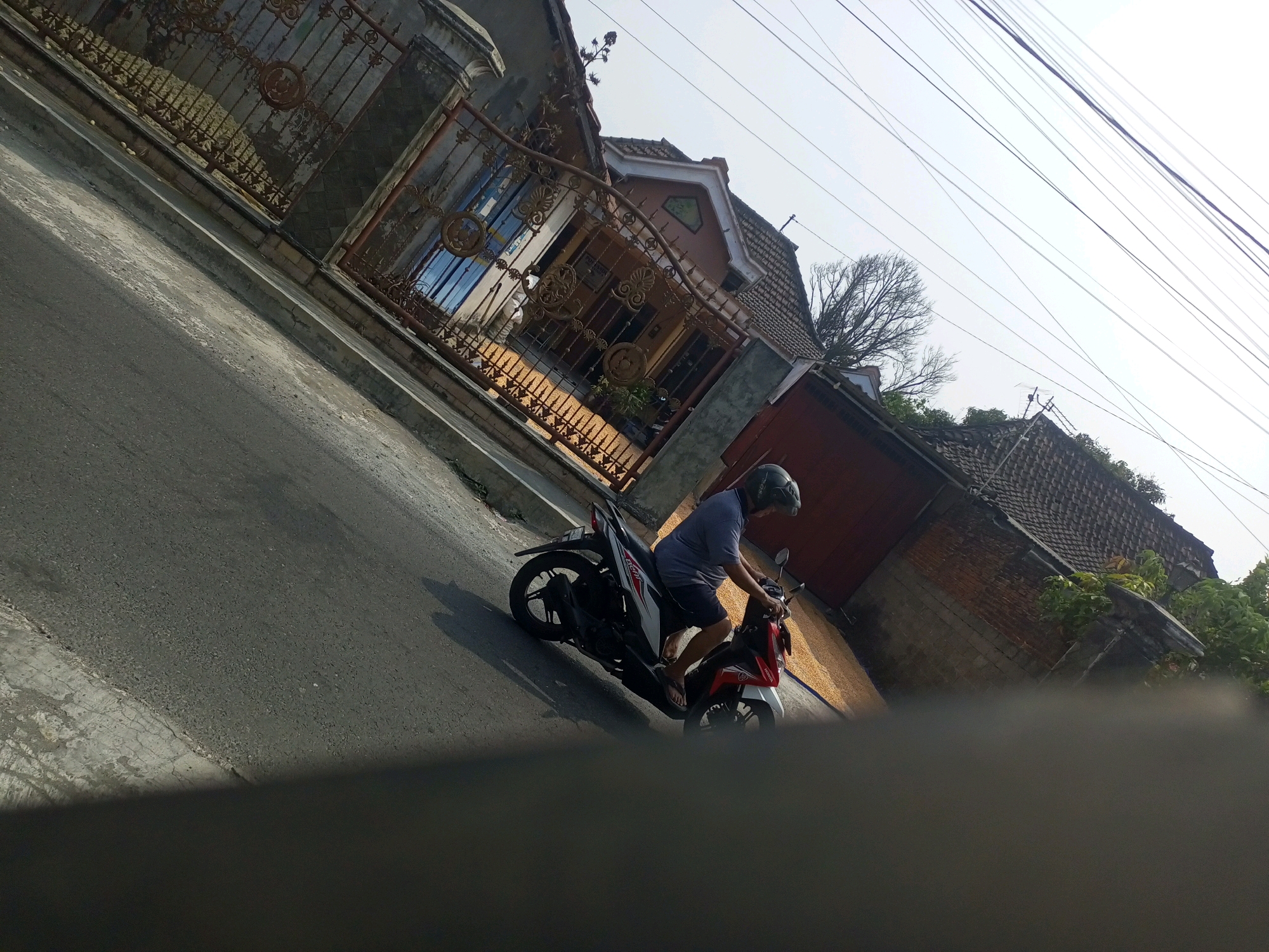 In Ngasem, Indonesia, a person wearing a dark helmet and a blue t-shirt is riding a white and red motorcycle down a paved road. The road is lined with a concrete sidewalk and a weathered brick building with a tiled roof. To the left of the road, an ornate metal gate with decorative scrollwork guards the entrance to a house with a brown-tiled roof and arched windows. Overhead, a tangle of power lines crisscrosses the bright, hazy sky. The scene is captured from a low angle, with a shadow cast on the lower part of the frame.