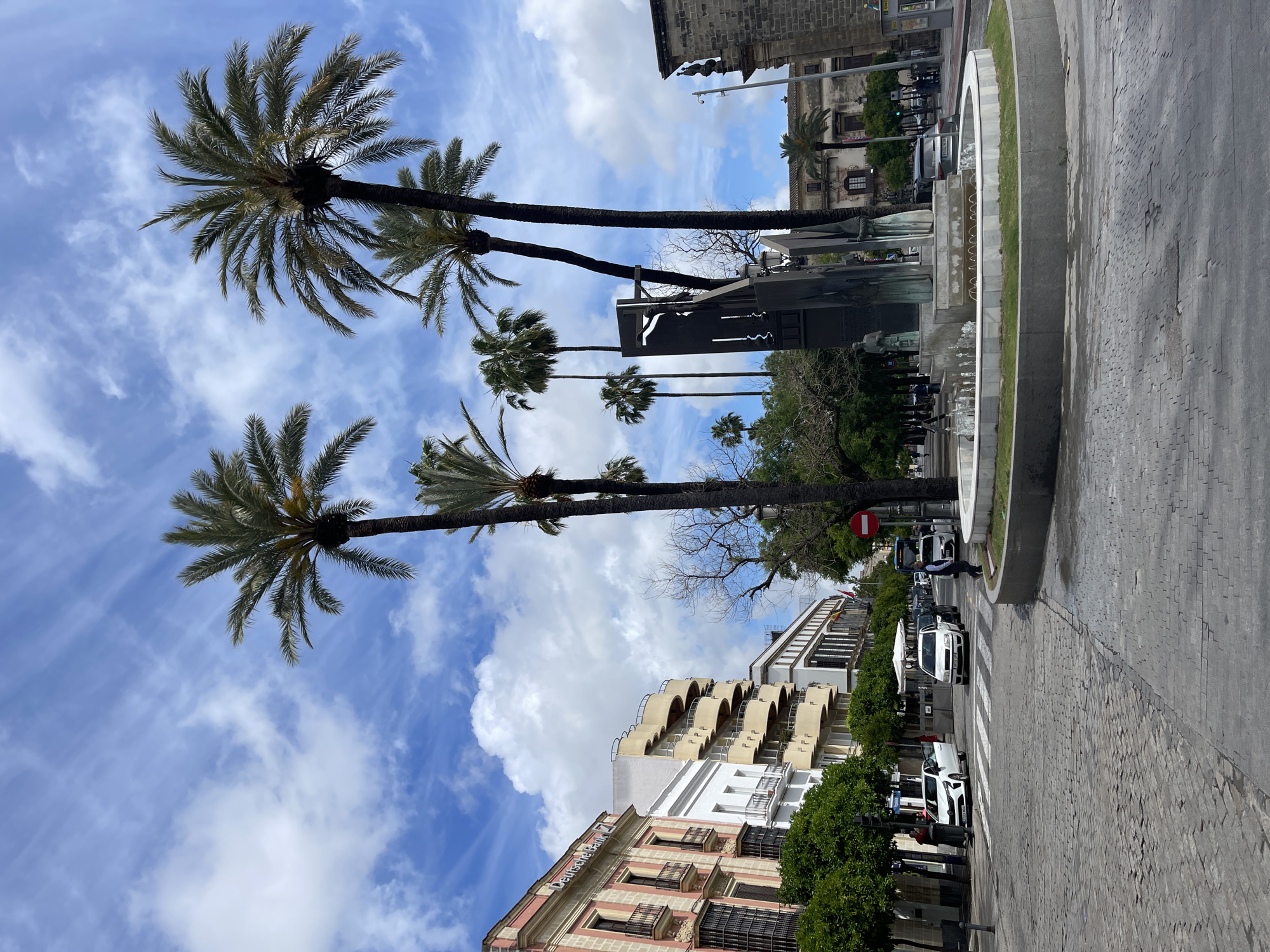 This street scene in Jerez De La Frontera, Spain, features palm trees lining a thoroughfare on a sunny day with a blue sky marked with puffy white clouds. The street is paved with cobblestones and includes a zebra crosswalk where a man is crossing. Adjacent to the crosswalk, cars and other vehicles are parked along the side of the road. Buildings line the side of the street, including a multistory building with arched balconies. In the center of the street, a modern sculpture stands next to a tiered fountain with a green border. A 'Do Not Enter' sign is visible at the intersection of the fountain and the street. Several other people can be observed in the background near the fountain and buildings.
