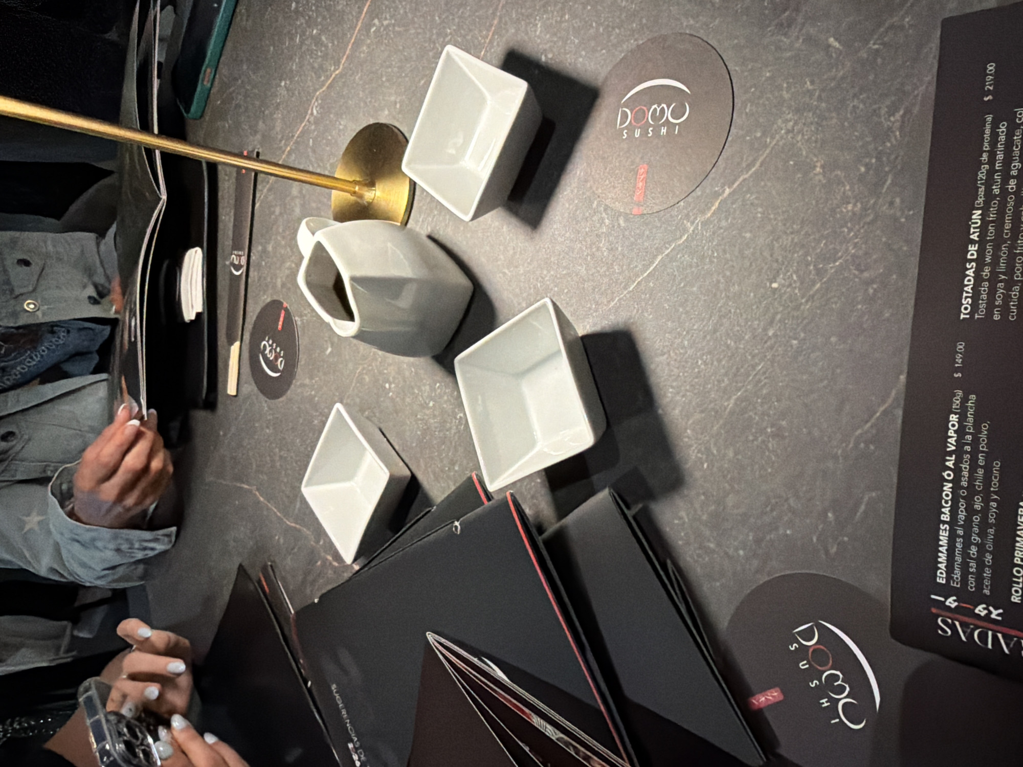 The image shows a table setting at Domo Sushi, a restaurant. The table is a dark, marbled surface, and there are several items arranged on it. There are white, square bowls, and a small pitcher on the table. Black menus with red accents are present. There are Domo Sushi branded coasters. Parts of people are visible including their hands. The OCR present suggests this is in San Bernardino Tlaxcalancingo, Mexico.