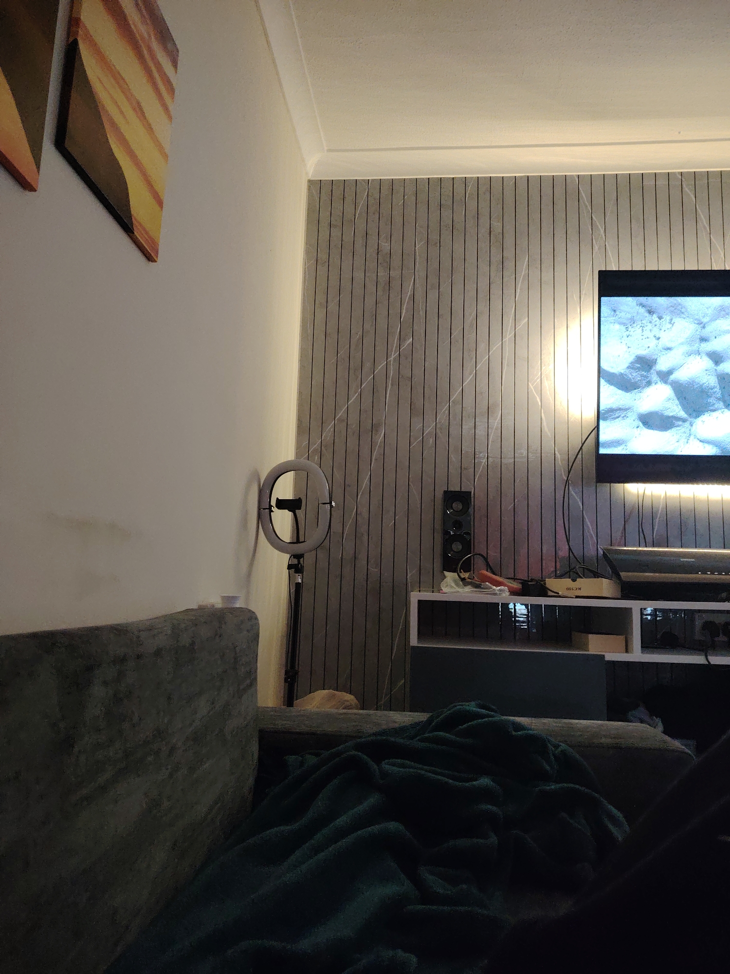 This image depicts an indoor domestic living space, specifically in Huntingdonshire, United Kingdom, captured from a low perspective, likely from a sofa in a dimly lit room. In the immediate foreground, to the left, is the dark grey, textured backrest and armrest of a sofa. A dark teal or deep green plush blanket is crumpled and draped over the sofa's seat and armrest to the right. The left wall is painted a light, off-white color. Two rectangular art pieces are mounted on this wall; the one on the right is more visible, featuring abstract, diagonal bands of warm colors including yellow, orange, and brown. Below these artworks, faint, irregular dark smudges are visible on the wall. A small, white, cylindrical object, possibly a cup, sits on an unseen surface to the right of the sofa. Further into the midground, positioned in the corner where the light wall meets an accent wall, is an illuminated LED ring light on a black tripod stand. A smartphone holder is visible within the ring. The ring light casts a prominent, elongated shadow on the light-colored wall to its left. The accent wall, starting from the corner and extending to the right, is covered with vertical panels or slats featuring a light grey base with distinct white, marble-like veining. On this accent wall, a large, black flat-screen television is mounted. The television screen is active, displaying an image composed of bright, light blue and white irregular, rounded shapes, resembling ice formations or abstract textures, under strong illumination. A thin, warm-toned LED strip light is affixed horizontally behind the television, casting an upward glow onto the accent wall beneath it. A thick black cable descends from the television. Beneath the television, a modern, wall-mounted entertainment unit is present, featuring a light-colored (likely white or very light grey) top shelf and darker (dark grey or black) lower sections. On the left side of this top shelf, a black, vertical speaker with two visible circular drivers is placed. Towards the center, several electronic items and cables are visible, including a rectangular beige box labeled "KE 100" and a coiled red-orange cable. A clear plastic bag with indiscernible contents rests nearby. Directly beneath the TV, on the shelf, is a dark-colored soundbar or media player. Below the main shelf, power outlets and additional cables are visible against the darker section of the unit. The ceiling is light-colored and features