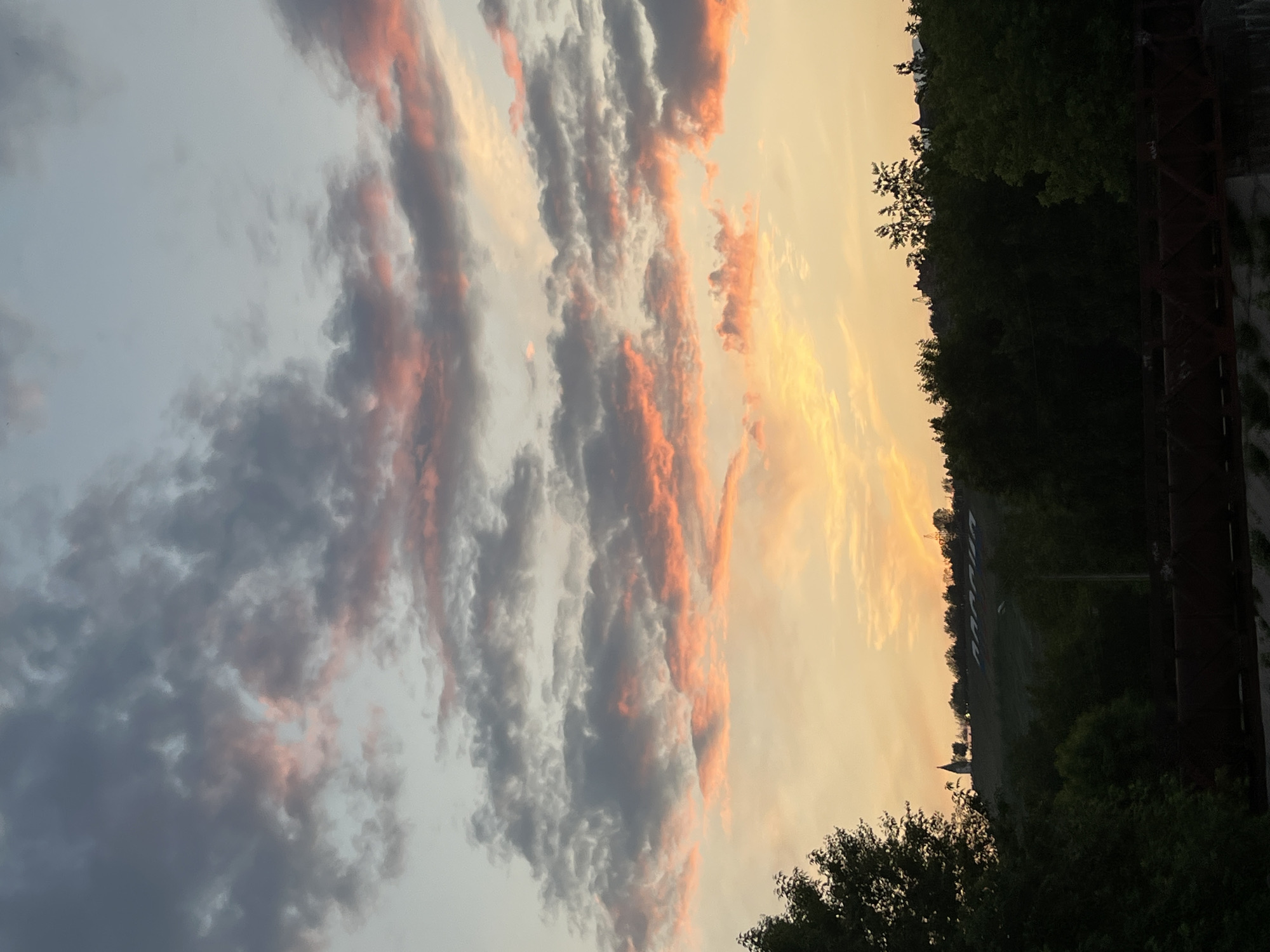 The image depicts a sunset over Krasnoyarsk, Russia, with a prominent church spire visible on a hill in the distance, accompanied by a hillside inscription. The sky is filled with dramatic clouds, tinged with vibrant hues of orange, pink, and gray, transitioning into a pale blue at the top. Silhouetted against the colorful sky is a line of trees and distant structures. In the foreground, dark green foliage and a rust-colored, industrial-looking structure, possibly a bridge, are visible. The overall scene captures a moment of natural beauty with a hint of urban landscape.