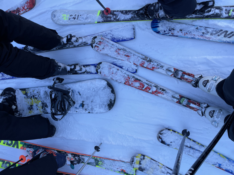 The image, captured from an overhead perspective, showcases a collection of winter sports equipment and parts of individuals on a snow-covered surface, likely on a mountain in Courmayeur, Italy. The scene primarily features skis, snowboards, ski boots, and ski poles, all partially covered in fresh snow.
Multiple people are implied, with their legs clad in black snow pants, and their feet secured in various bindings. In the upper left, a pair of black boots are attached to skis. Another pair of skis prominently displays the brand "FISCHER" and model details such as "RC4" or "RCR," featuring red, white, and black designs, with white ski boots visible. These skis are positioned near another pair of black ski poles and a partial view of a ski boot.
Toward the center, a black snowboard with black bindings, also dusted with snow, is visible. It features yellow and black graphics, with some indistinct text that could be "STK" or "SOL". Nearby, the lower legs and boots of a person using this snowboard are visible. Another set of skis with yellow, black, and white patterns can be seen in the lower left, accompanied by ski poles. At the top of the frame, another pair of skis shows partial text "4F" and potentially "HEAD."
The entire setting is dominated by bright white snow, indicating cold weather and potentially recent snowfall. The even lighting suggests it is daytime, possibly under an overcast sky, as there are no distinct shadows. The arrangement of the gear suggests a group has gathered, perhaps taking a break or waiting, during their skiing and snowboarding activities on a resort slope.