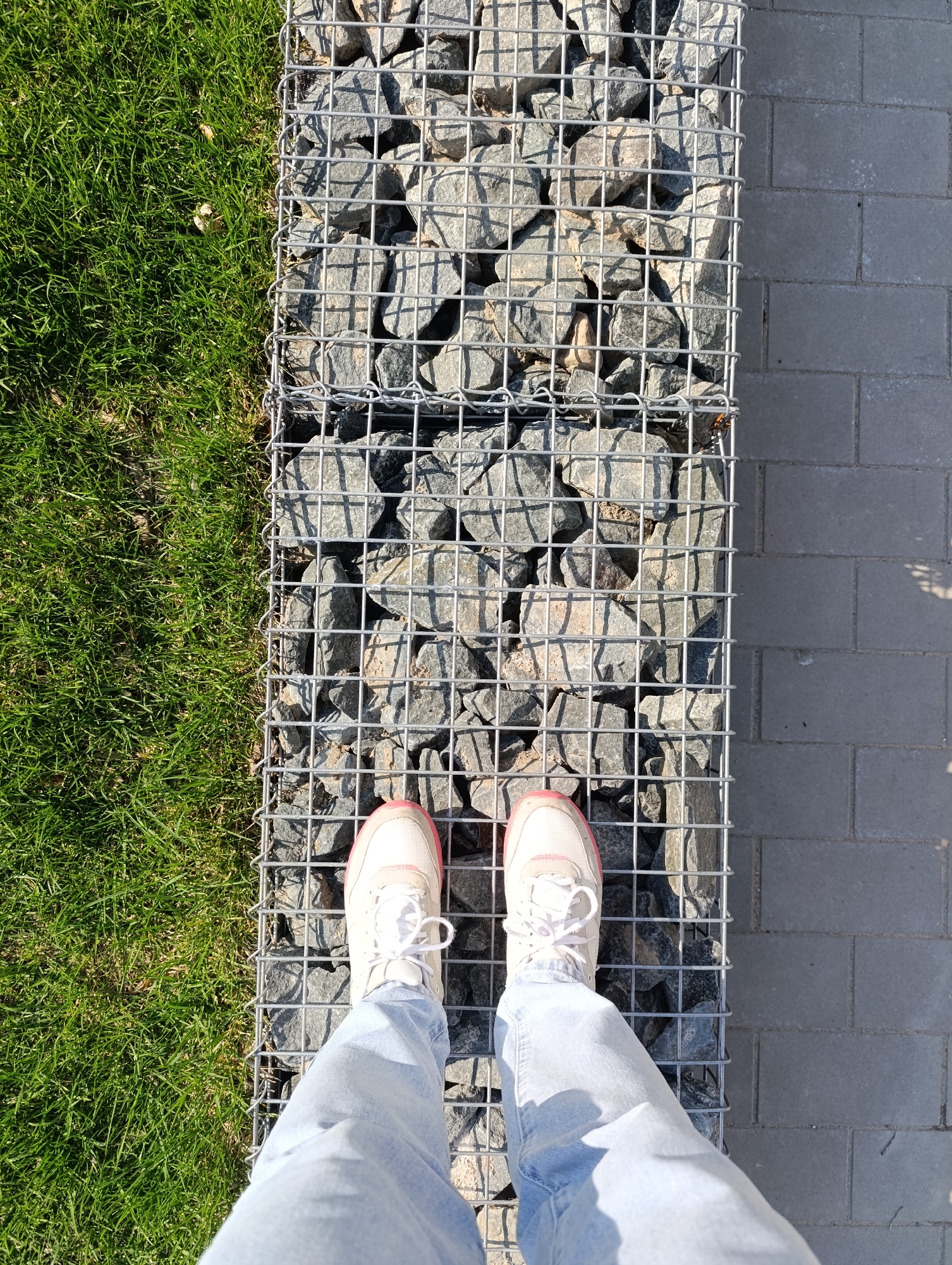 A person stands on a gabion wall, which is constructed from wire cages filled with stones. The shoes of the person are white with pink trim. The person is wearing light blue jeans. To the left of the gabion wall is a grassy area. To the right of the gabion wall is a paved walkway. The location is Unknown, Unknown.
