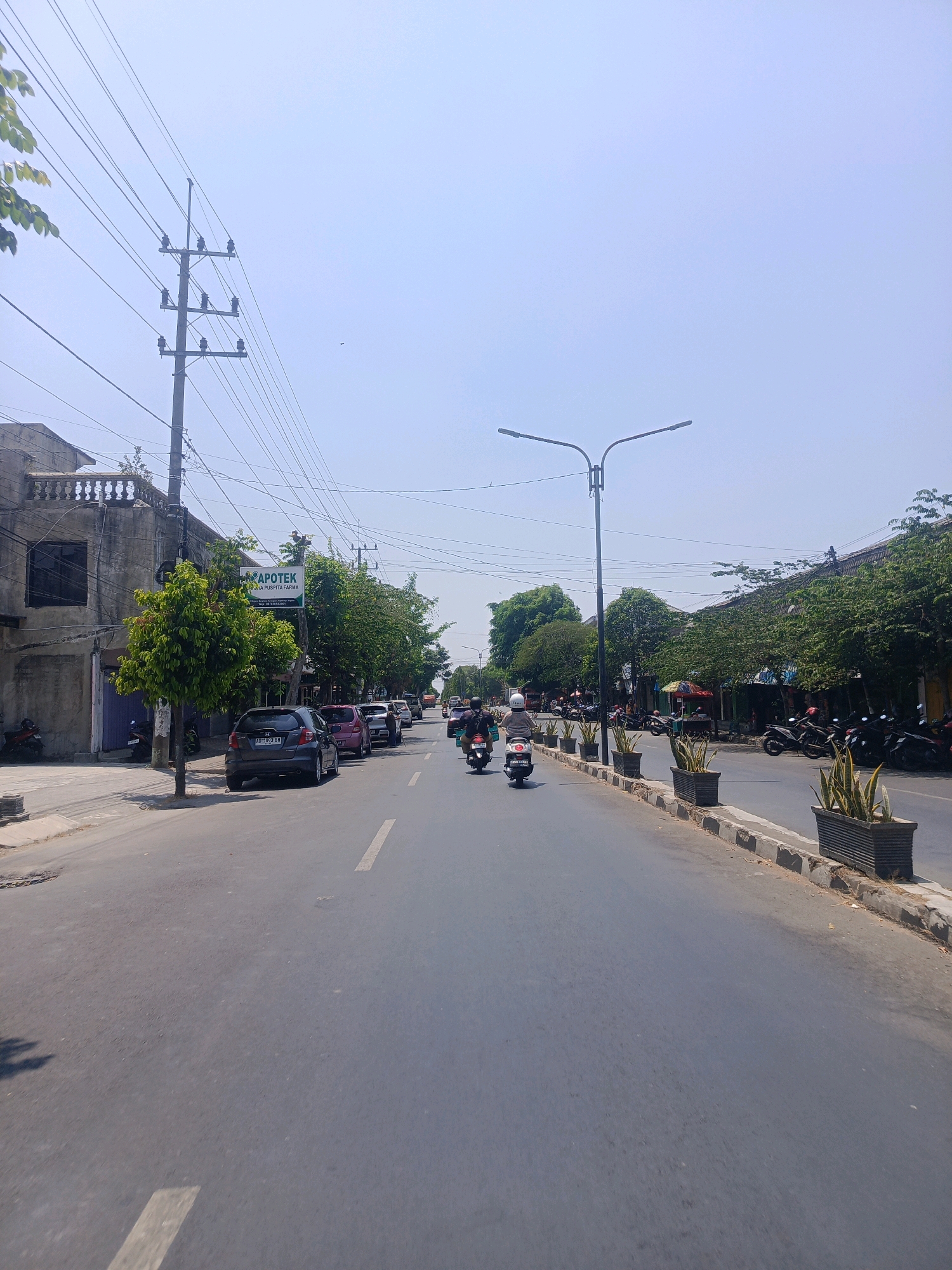 The image captures a street scene in Sukowinangun, Indonesia, under a clear, bright sky. The perspective is from a moving vehicle, looking down a two-lane road lined with buildings and trees. On the left, a two-story building with a textured facade and balconies is visible, with a motorcycle parked in front. Further down the street, a green sign for "APOTEK PUSPITA FARMA" is mounted on a pole, indicating a pharmacy. Several cars are parked along the left side of the road, including a dark gray hatchback with license plate "AD 1093 BH".
The right side of the road features a sidewalk with a row of rectangular planters containing spiky green plants. Beyond the sidewalk are buildings with tiled roofs and awnings, with parked motorcycles visible. Several people are riding motorcycles on the road, some wearing helmets. In the distance, the road curves slightly, and more trees and buildings are visible. A street lamp with two lights is positioned on the right side of the road. Overhead, a complex network of electrical wires runs across the sky, supported by utility poles. The overall impression is of a typical urban street on a sunny day.