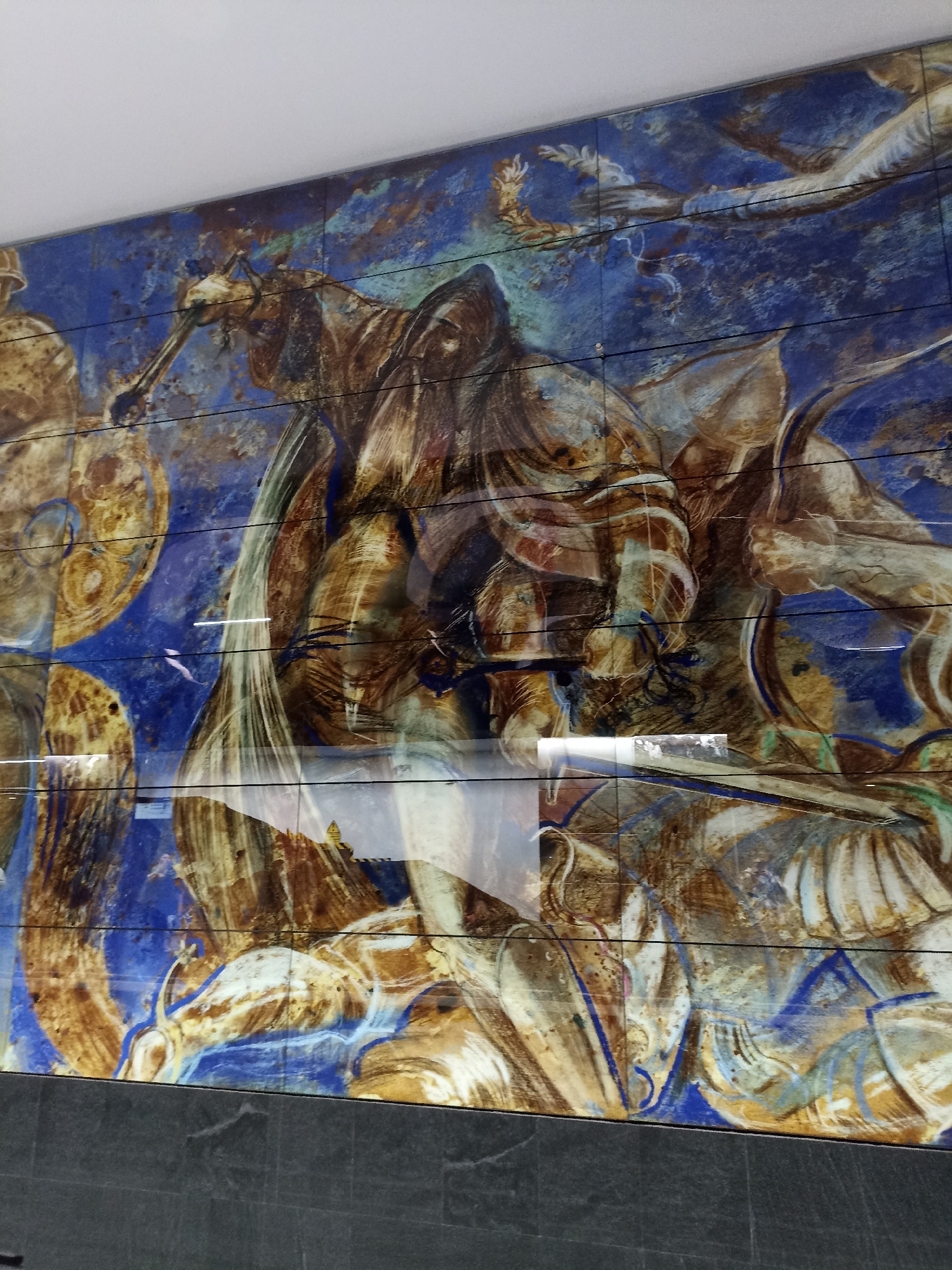 The image shows a close-up view of a large, colorful mural or mosaic adorning a wall. The artwork features abstract, dynamic forms in shades of blue, brown, and gold. It appears to depict figures, possibly mythological or allegorical, in a dramatic and energetic composition. The artwork is composed of square tiles and is mounted on a wall above a dark, textured base. Reflections are visible on the surface of the artwork, indicating it might be protected by glass or a similar material, possibly within an indoor public space. The artwork is located in the Moscow Metro, specifically on the platform of the Komsomolskaya station.