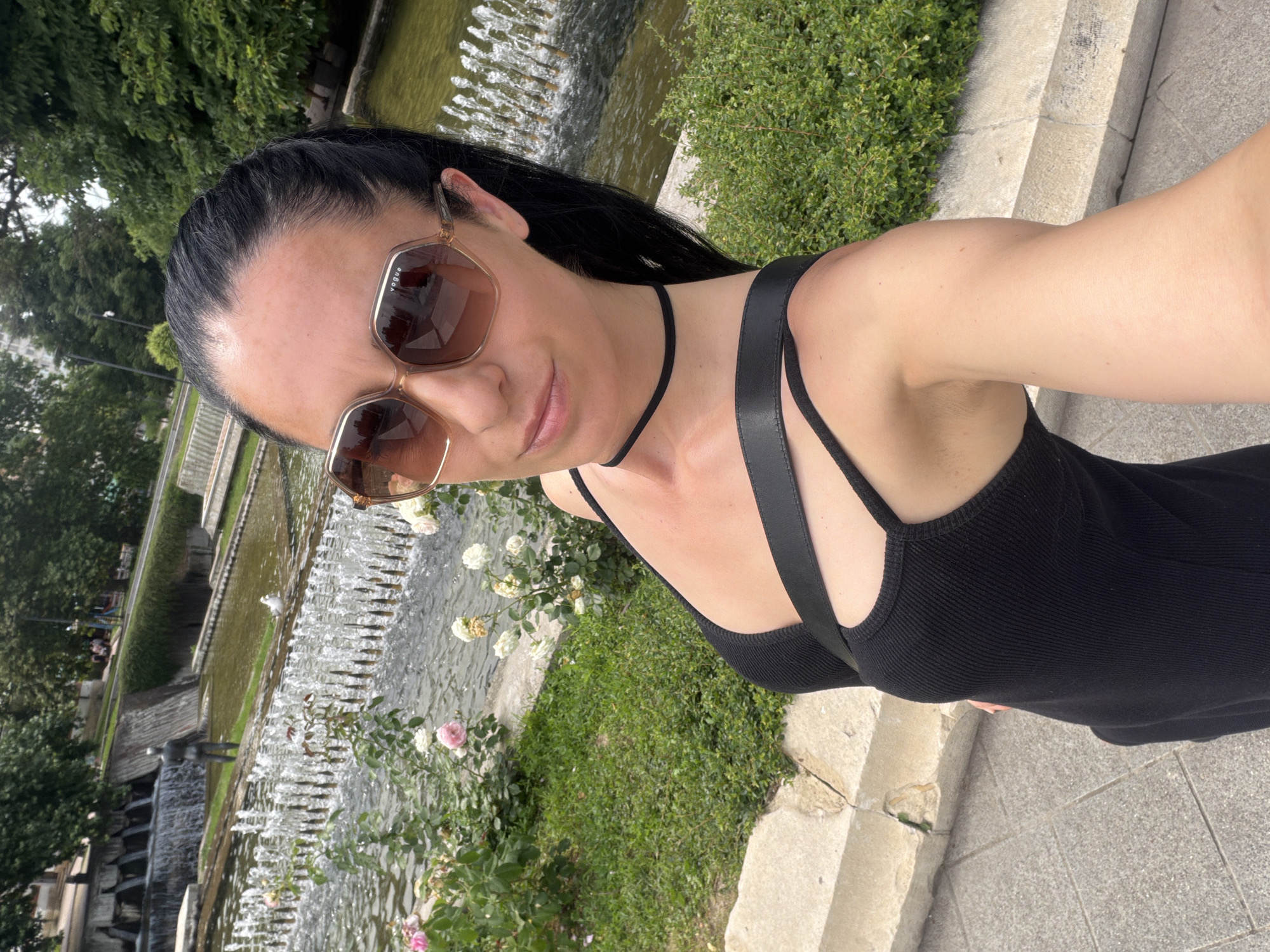 A woman takes a selfie in front of a fountain in a park, with a statue visible in the background. The woman is wearing a black dress and sunglasses. The fountain has several rows of water jets, and there are rose bushes and greenery surrounding it. The statue appears to be a male figure standing in a shallow pool of water. The scene is set in a park with trees and a paved walkway. This location is the central fountain in the city of Pleven, Bulgaria, known as the "Fountain of Youth."