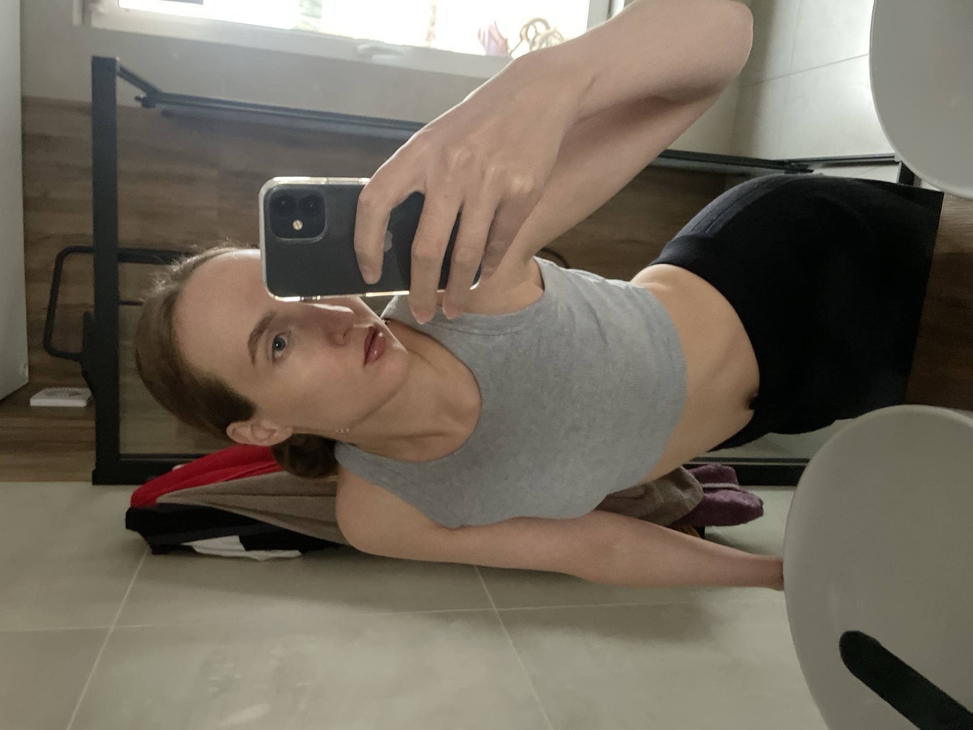 A woman is lying on a tiled floor in a bathroom, taking a selfie with a black smartphone. She is wearing a gray tank top and black shorts. Clutter, including clothing and towels, is scattered around her. A toilet is partially visible in the foreground. The location is a bathroom. The city is not discernible from the image.