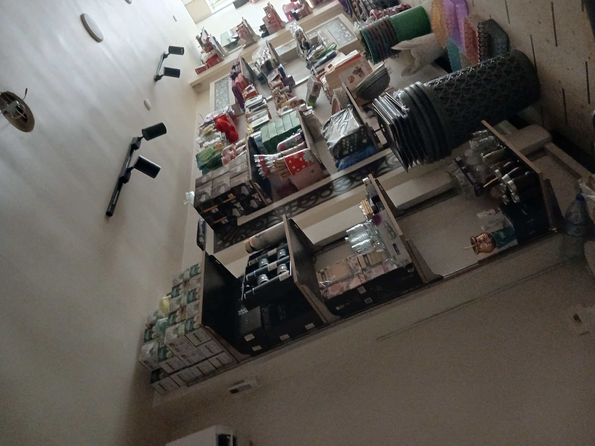 The image depicts the interior of a shop, likely a variety store or a small market, located in Rukn ad-Din Municipality, Syria. The shop is filled with shelves stocked with various consumer goods. On the shelves, one can observe stacks of boxed products, possibly food items or household goods, some with green and white packaging. Other items on display include what appear to be kitchenware, such as cups, bottles, and containers, along with decorative items and possibly stationery. In the foreground, to the right, a large stack of dark-colored, patterned plastic containers or bins is visible, alongside smaller colorful plastic trays or containers. The lighting in the shop appears to be artificial, with track lighting fixtures visible on the ceiling. The overall impression is one of a densely stocked retail space.