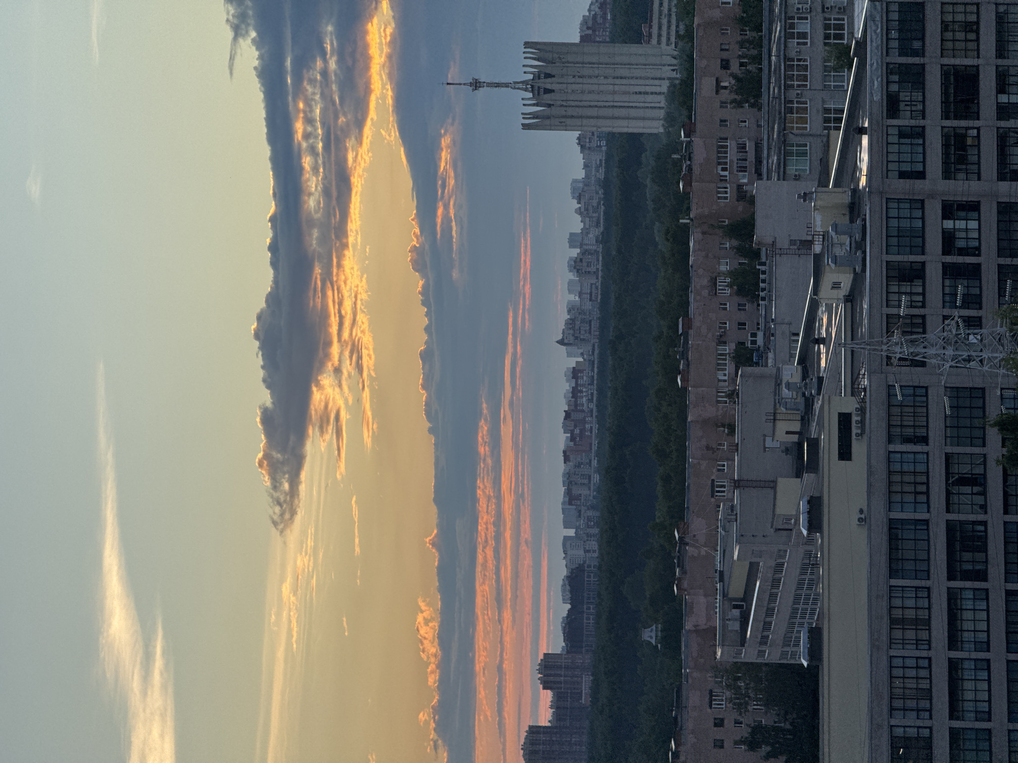 The image shows a city skyline at dusk. The sky is filled with dramatic clouds, tinged with orange and yellow from the setting sun. In the foreground, a series of multi-story industrial or residential buildings are visible, their rooftops covered with vents and pipes. A distinctive, angular tower with a spire stands out on the right side of the image, set against the backdrop of a dense green forest that separates the closer buildings from the distant cityscape. The distant skyline is characterized by numerous apartment blocks and other structures. The overall impression is an urban environment transitioning into evening, with the sky providing a vibrant and colorful spectacle.