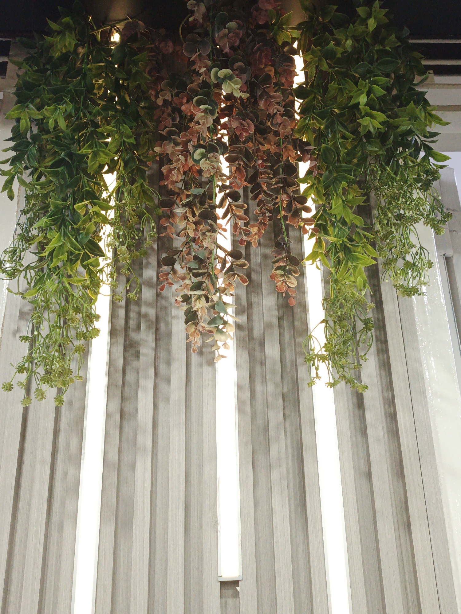 The image presents a view looking upwards at hanging faux greenery, featuring strands of green and brownish-red artificial plants. These artificial plants are suspended from above, obstructing the view of the light source illuminating the scene. The background consists of vertical gray panels, interspersed with bright white light strips. No individuals, animals, or identifiable branded objects are present in the image. The environment appears to be an interior space with decorative elements. The specific location within the city of Tinaquillo, Venezuela, cannot be determined based on the image alone.