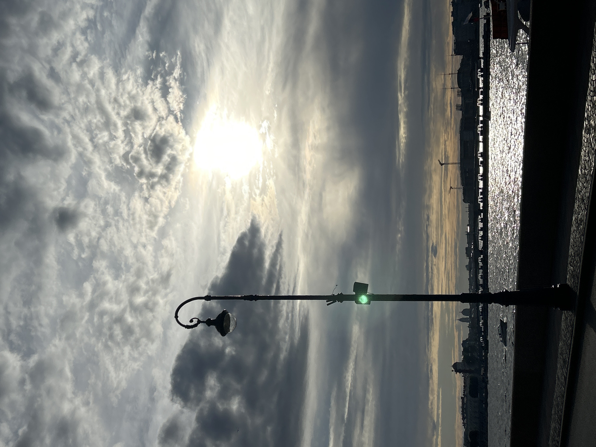 This is a vertical, low-angle, silhouette shot of a streetlamp against a dramatic sky. The streetlamp, a dark, ornate object, is positioned in the foreground, its curved arm and single fixture visible. To the right of the lamp, a green traffic light glows, casting a small, vibrant spot of color against the muted tones of the sky.
The sky itself is a dominant feature, filled with a mix of clouds. Wispy, white clouds are illuminated by a bright, hazy sun, which is partially obscured but still intensely luminous. Darker, heavier clouds streak across the frame, creating a sense of depth and contrast. The overall impression of the sky is one of dynamism and perhaps impending change.
In the background, a body of water shimmers under the sunlight, reflecting the sky's brilliance. Beyond the water, a city skyline is visible in silhouette. Buildings, including domes and spires, create a jagged line against the horizon. A few boats or ships are faintly discernible on the water.
The scene appears to be set outdoors, likely near a waterfront in an urban area. The time of day is difficult to pinpoint precisely due to the strong backlighting, but the quality of light suggests it could be late afternoon or early morning, when the sun is lower in the sky. The weather seems to be a mix of sun and clouds, creating a moody atmosphere. There is no visible text in the image.
