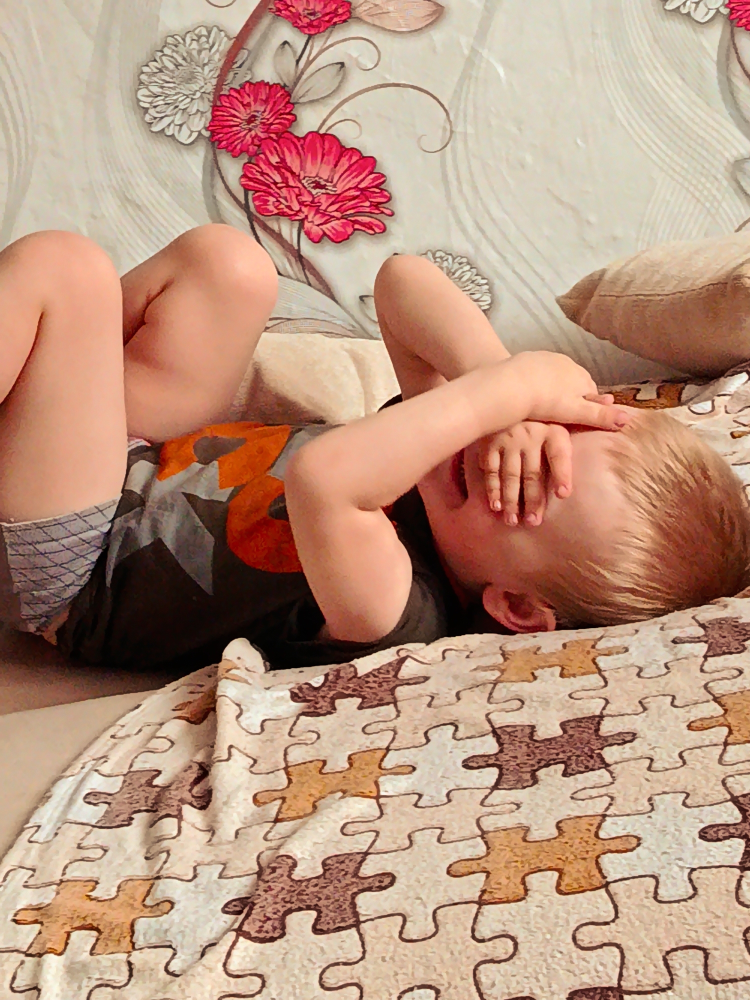 A young child with light-colored hair is depicted lying on their back on a bed in an indoor setting. The child's knees are bent, and their hands are covering their face, obscuring their eyes and nose. They are dressed in a dark t-shirt with an orange and grey graphic design, and grey patterned underwear or shorts are visible.
The bed is covered with a blanket featuring a distinct jigsaw puzzle pattern in shades of cream, tan, and brown. A beige pillow rests near the child's head on the right side of the frame. The wall behind the bed is adorned with wallpaper showcasing large floral designs, including prominent red and white flowers on a light background.
The lighting suggests it is daytime, although no direct light source is visible. The act of covering the face could indicate playfulness, shyness, or perhaps a moment of mild distress, but the child's precise emotion is not clearly discernible due to the obscured facial features. No explicit text is visible within the image. The scene conveys a moment of rest or play within a home environment, located in Фоки, Russia.