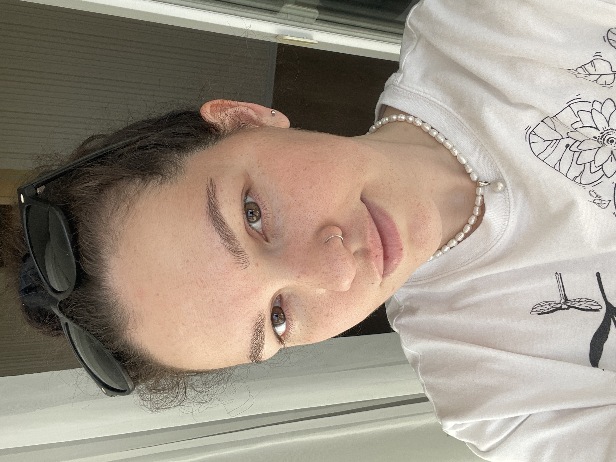 A close-up shot captures a woman with fair skin, brown eyes, and dark hair. She wears a white T-shirt with a black graphic design, a pearl necklace, and a small hoop nose ring. Sunglasses are perched on her head. She appears to be indoors. The location is unknown.