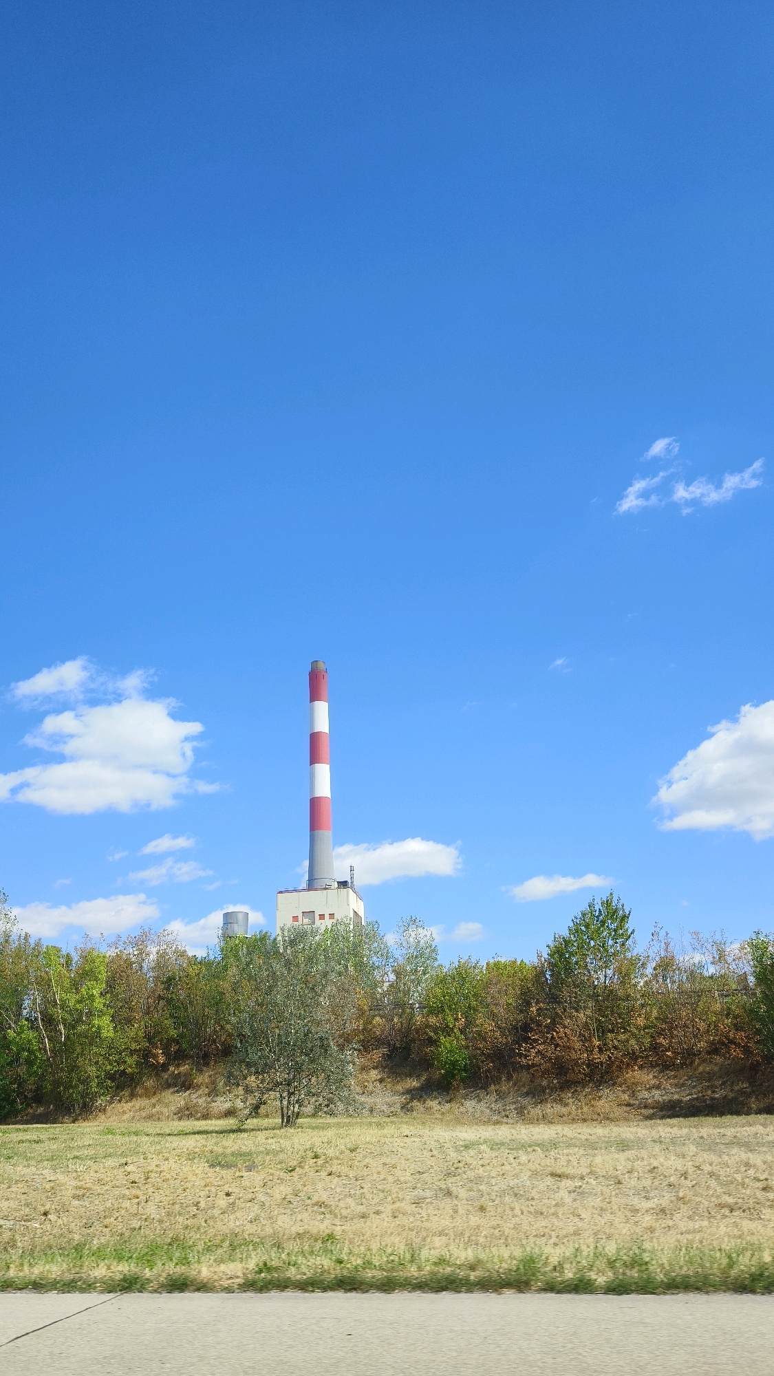 The image depicts a landscape dominated by a bright blue sky with scattered white clouds. In the middle ground, a tall industrial chimney with red and white stripes stands prominently against the sky. The chimney is attached to a white industrial building with several windows. A line of trees with varied foliage, ranging from green to brown, stretches across the middle of the frame, forming a natural barrier between the foreground and the industrial structures. The foreground consists of a dry, grassy field. The bottom of the image shows a section of a gray road or pathway. The scene appears to be located in Korneuburg, Austria, possibly near the Korneuburg Power Station, given the presence of the distinctive chimney. No people are visible in the image.