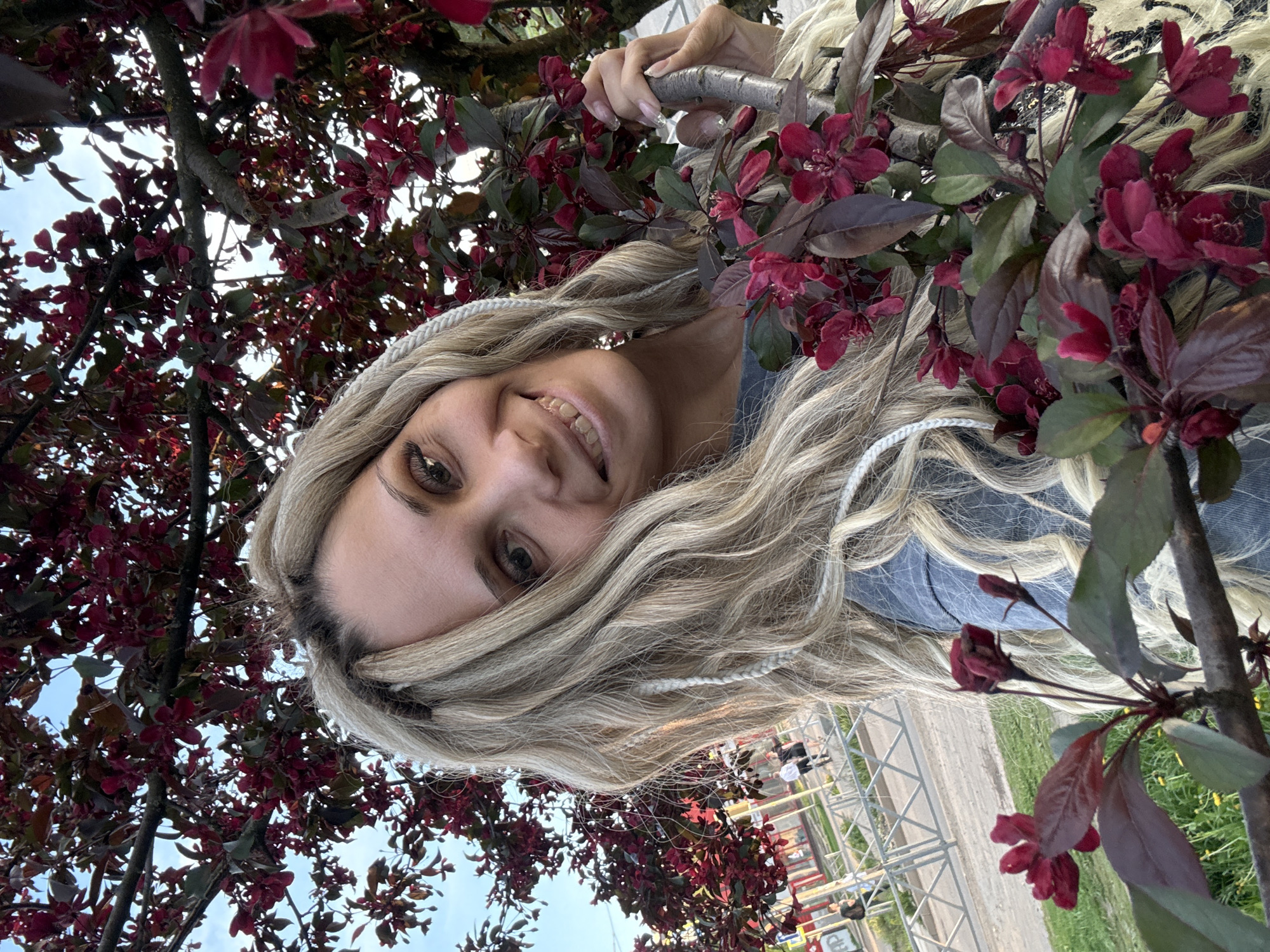 A young woman with blond hair and white braids is looking at the camera and smiling. She is holding onto a tree branch with dark red flowers. The background shows a park, possibly in the city of Rusanovo, Russia, with a paved area and people in the distance. A metal structure, possibly a fence or railing, is also visible.