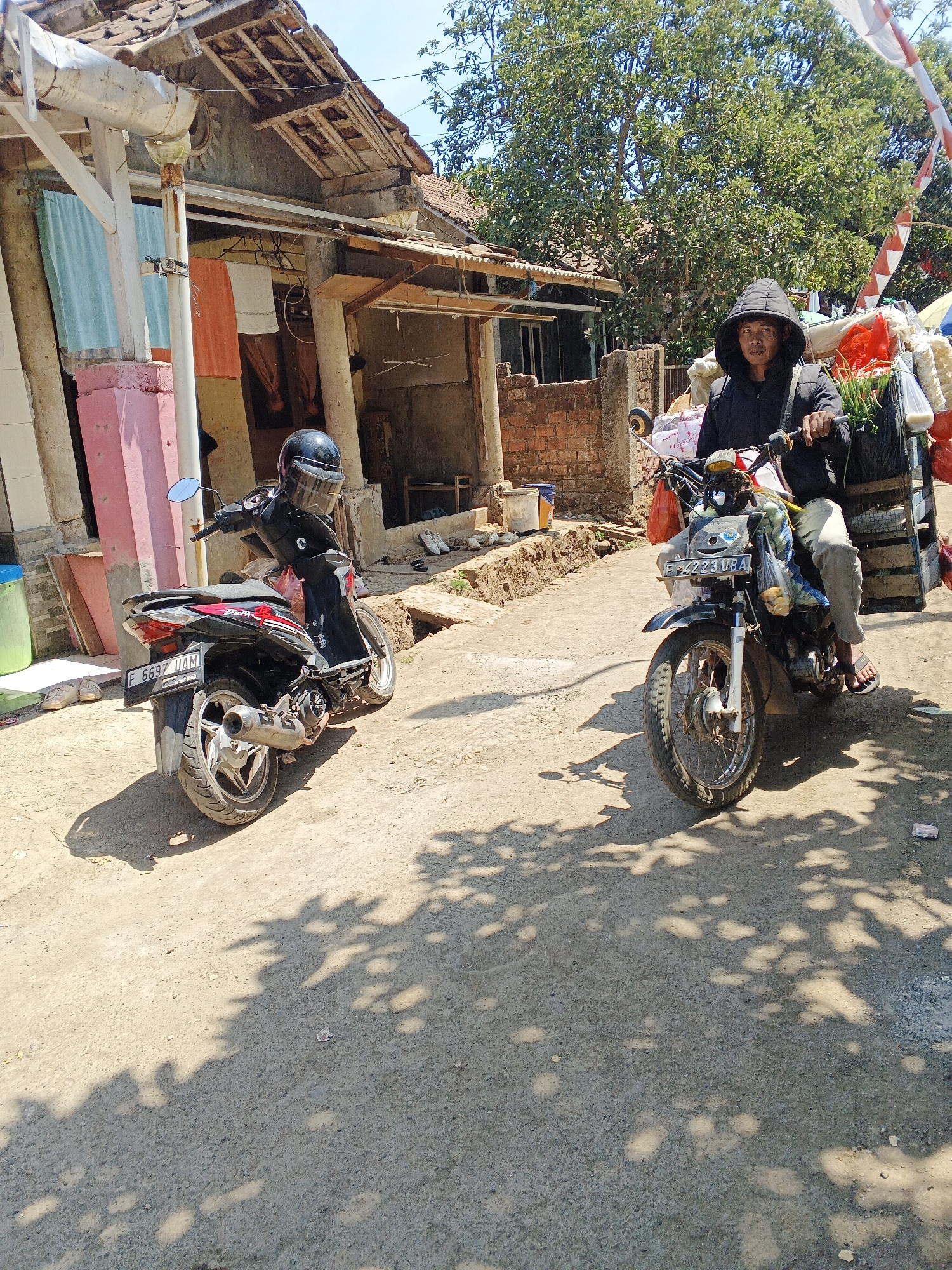 A man wearing a black hooded jacket and gray pants rides a motorcycle with a cargo rack on the back. He is in a dirt road in Sukaharja, Indonesia, with a house made of brick and wood on the left side of the image and trees in the background. The motorcycle has license plate F 4223 UBA. Another motorcycle, a black scooter with license plate F 6697 UAM, is parked to the left of the man. The scooter has a black helmet on its seat. Laundry hangs from the house on the left, and the scene is lit by sunlight, casting shadows on the ground.