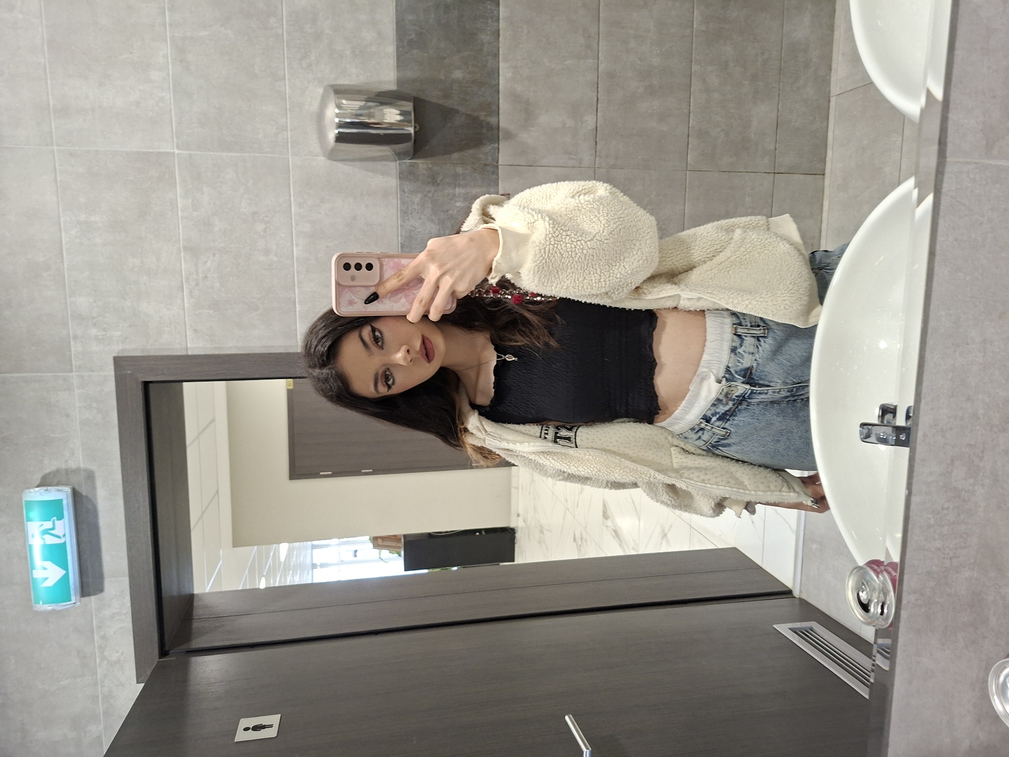 A young woman with long dark hair is taking a selfie in a bathroom. She is wearing a black crop top, blue jeans, and a light-colored, fuzzy jacket. The bathroom has grey tiled walls and floors, a metal hand dryer, and a white sink. An exit sign is visible on the wall near the mirror. The location appears to be a public restroom in an indoor setting, likely in Sofia, Bulgaria.