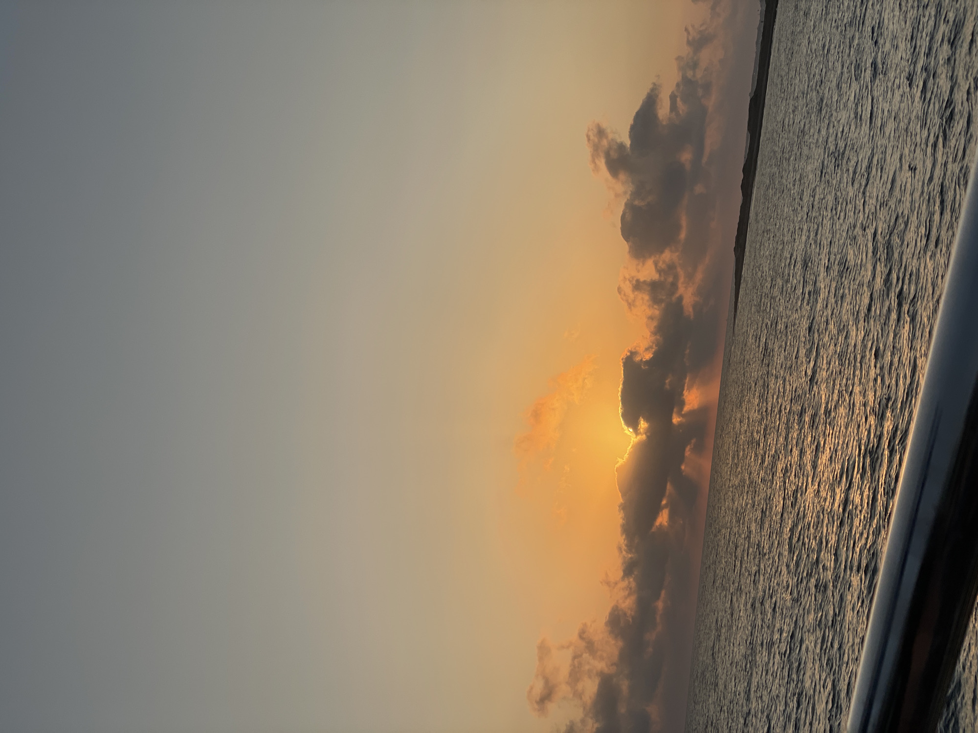 The image captures a serene sunset over the Mediterranean Sea, likely near Yeroskipou, Cyprus, as indicated by the specific location context. The sky transitions from a pale blue at the top to a warm orange glow near the horizon, where the sun is partially obscured by dark, dramatic clouds. Rays of sunlight pierce through the clouds, creating a beautiful crepuscular effect. The sea is calm, with gentle ripples reflecting the colors of the sky. In the foreground, a metallic railing, possibly from a boat or a pier, runs horizontally across the bottom of the frame, adding a sense of depth and perspective. The distant coastline appears as a dark, low-lying strip against the horizon. There are no people or explicit human activity visible in the scene.