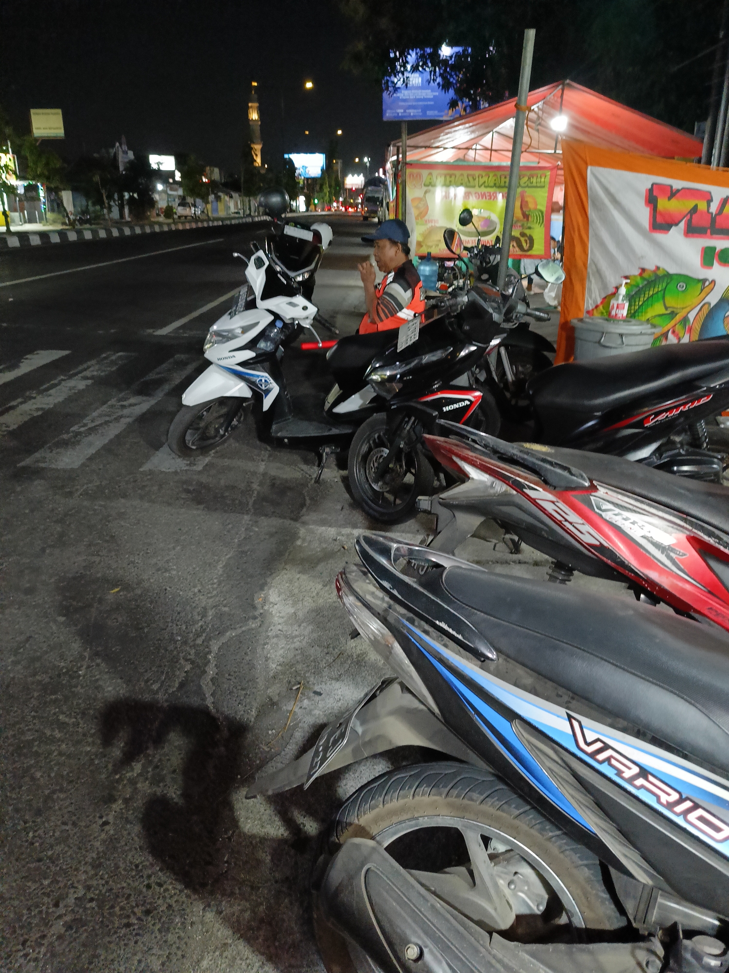 The image captures a nighttime scene along a city street in Kebondalem Kidul, Indonesia. The foreground is dominated by several parked motorcycles. A white Honda Vario is positioned prominently on the left, its front wheel turned slightly. To its right and slightly behind, a black Honda Vario is parked. Further back, another motorcycle, possibly a red and white Vario, is visible. A person wearing a blue cap and an orange and white striped shirt is seated on one of the motorcycles, facing left and appearing to be eating or smoking. They are wearing a dark blue cap. Behind the motorcycles, a temporary stall with an orange and red tent is set up, displaying various banners with text and images, including a depiction of a fish. A cylindrical trash bin is situated near the stall. Across the street, a road is visible with a pedestrian crossing marked by white stripes. Streetlights illuminate the area, and in the background, a tall minaret of a mosque stands against the dark night sky. The overall environment suggests a street-side establishment operating at night.