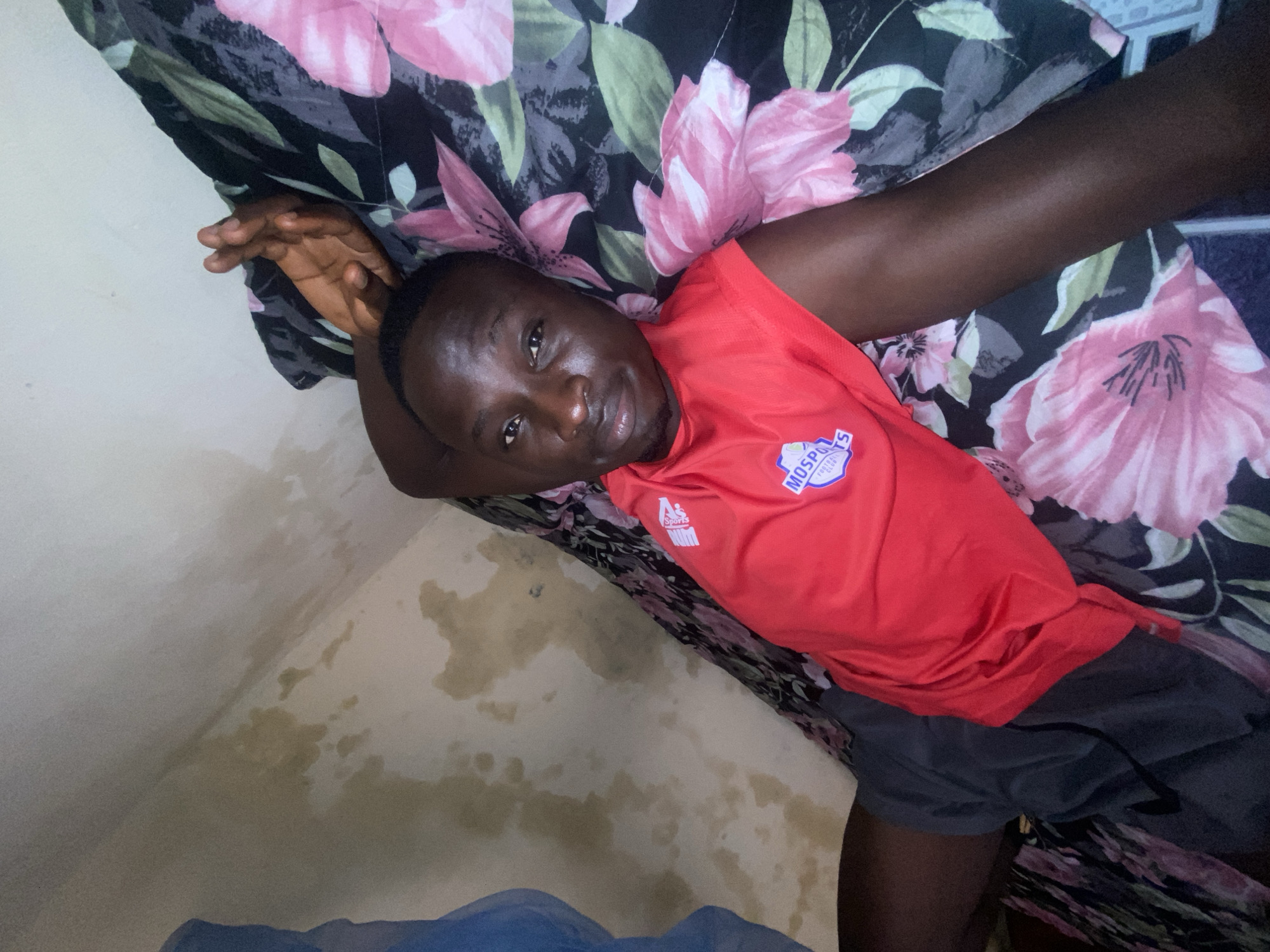 A dark-skinned male is lying on a bed with a floral patterned comforter. He is wearing a red sleeveless shirt with a white logo and grey shorts. His right arm is bent at the elbow, with his hand resting on the pillow. The background shows a wall with water stains. The lighting is dim, suggesting an indoor setting at night or in a poorly lit room. The overall scene appears to be a bedroom in a residential area in Barden Yana, Nigeria.
