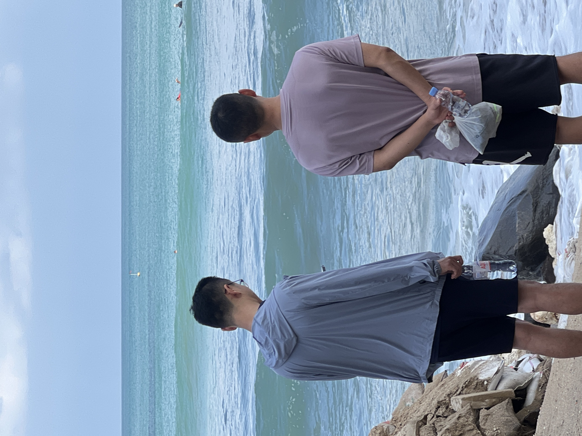 Two individuals stand on a rocky beach facing the ocean in Kuta, Indonesia. The person on the left wears a grey hooded jacket and black shorts, holding a bottle of water. The person on the right wears a light purple t-shirt and black shorts, with their hands clasped behind their back, holding a plastic bag containing a water bottle and some debris. The ocean is a vibrant turquoise with moderate waves, and several people are visible in the water, some swimming and others on surfboards. The sky is overcast, with a cloudy horizon. The foreground is composed of rocks, sand, and some discarded materials.