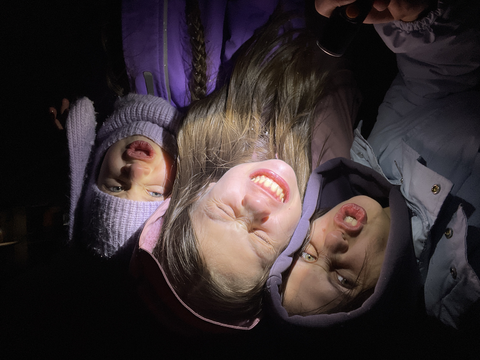 Three young individuals are positioned closely together, presumably outside, under a concentrated light source in the darkness. The individual on the left wears a lilac-colored knit hat and has a partially obscured face. The central individual is smiling widely, revealing teeth, and appears to be partially squinting under the light. The individual on the right is wearing a hooded jacket and making a distorted facial expression. A hand holding a light source is visible at the top center of the frame. The setting is dark. The city, Pozim', Russia, cannot be inferred from the image.