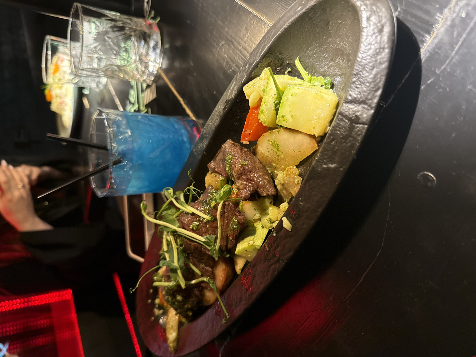 The image shows a plate of food and drinks on a table, with a person's hand partially visible in the background. The plate contains cooked meat and a variety of vegetables. A blue drink with straws is also visible next to the plate. There are glasses behind the plate as well. The setting appears to be a restaurant or bar with dim lighting. There is no information available in the image to confirm that it is located in Saint Petersburg, Russia.