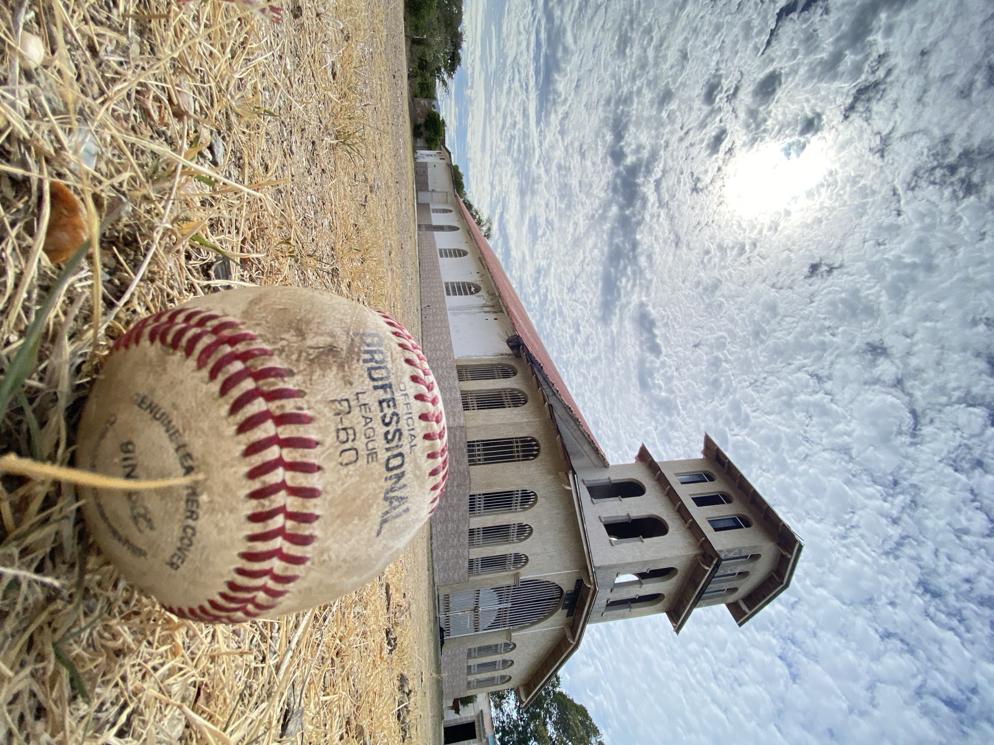 The image presents a close-up, angled shot of a baseball resting on dry grass, with a building and sky in the background. The baseball is an "Official Professional League R-60" ball, showing signs of wear. Behind the baseball, a long, low building with a tower is visible. The building has numerous arched windows with bars. The sky is filled with white, puffy clouds. The location is Juan Griego, Venezuela.