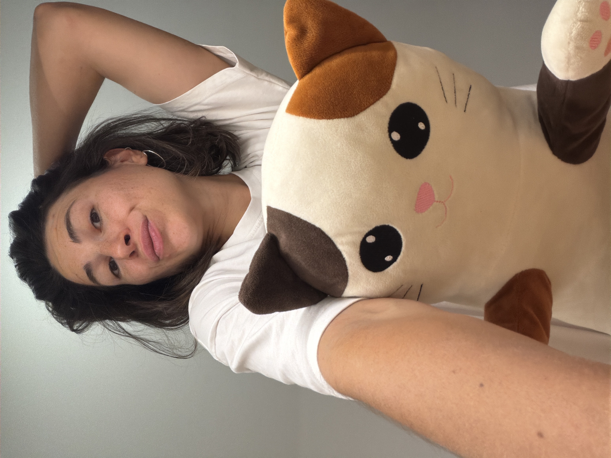 A woman with brown hair, wearing a white T-shirt and hoop earrings, is lying on her side holding a large plush cat toy with brown and white fur. The toy has embroidered black eyes and a pink nose and mouth. The background is a plain, light gray wall. There is no indication of the location being in Budva, Montenegro.