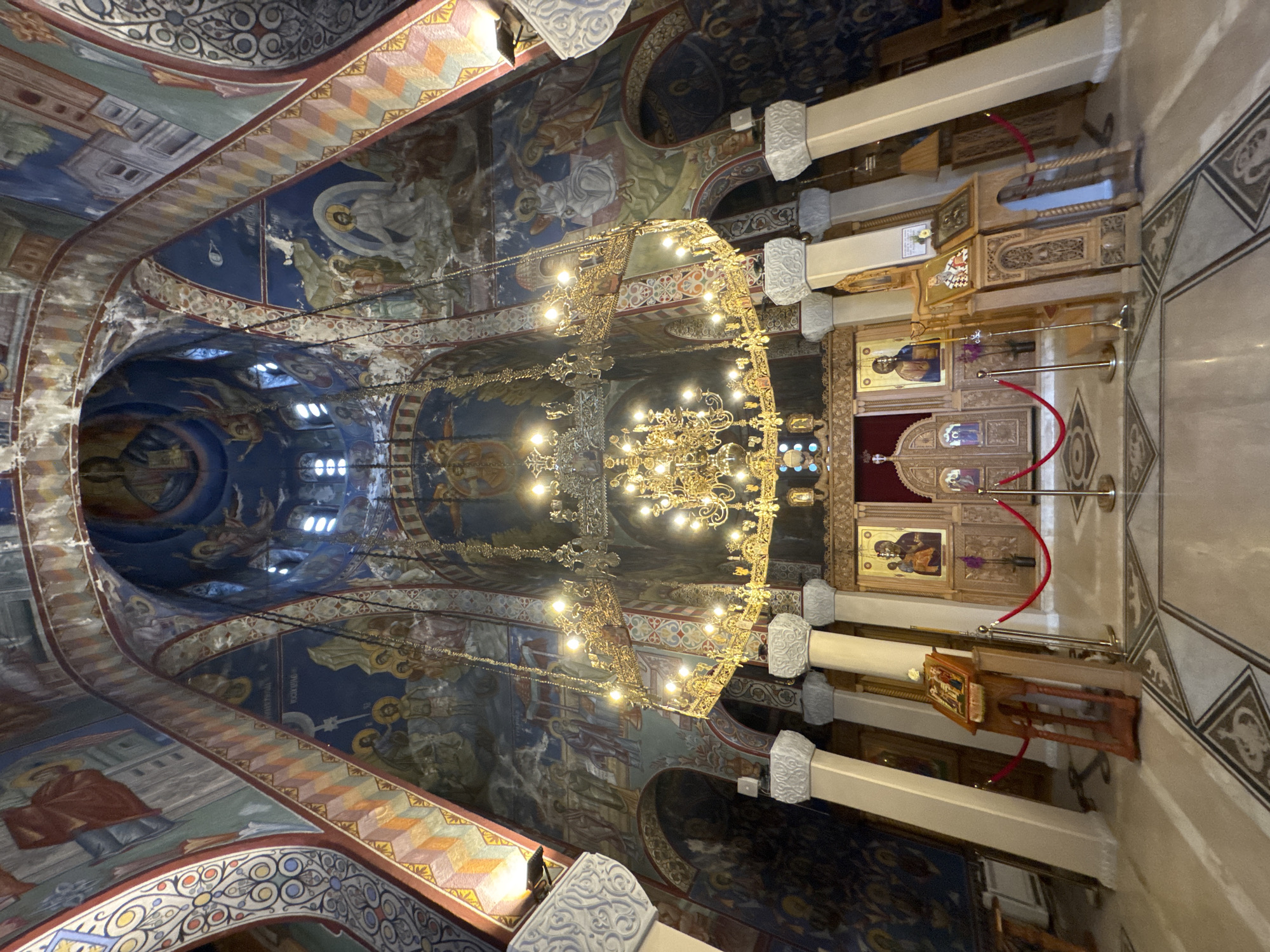 The image shows the interior of a grand Orthodox church, likely the Holy Transfiguration Cathedral in Trebinje, Bosnia and Herzegovina, given its ornate decoration and typical Orthodox church architecture. The vaulted ceiling and walls are covered in vibrant, detailed frescoes depicting religious scenes and figures. A large, ornate golden chandelier hangs prominently in the center of the space, illuminated by numerous bulbs. In the foreground, a marble floor with intricate geometric patterns is visible. The area in front of the altar features a wooden iconostasis with several religious icons, and a large wooden double door. Red velvet ropes on stanchions are used to demarcate areas, suggesting a space accessible to visitors but with restricted access to certain areas. White columns with decorative capitals support the ceiling. The lighting is a mix of natural light filtering through high windows and artificial light from the chandelier and wall sconces. No people are visible in the scene.