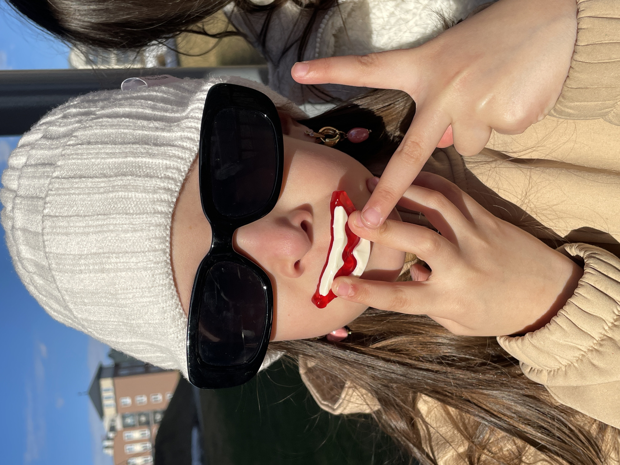 A young woman wearing a white knit hat and oversized black sunglasses is holding a red and white object near her face. She is making a peace sign with her hand. The background includes a building, a blue sky, and what appears to be an outdoor setting. The location is Pervomaysky, Russia.