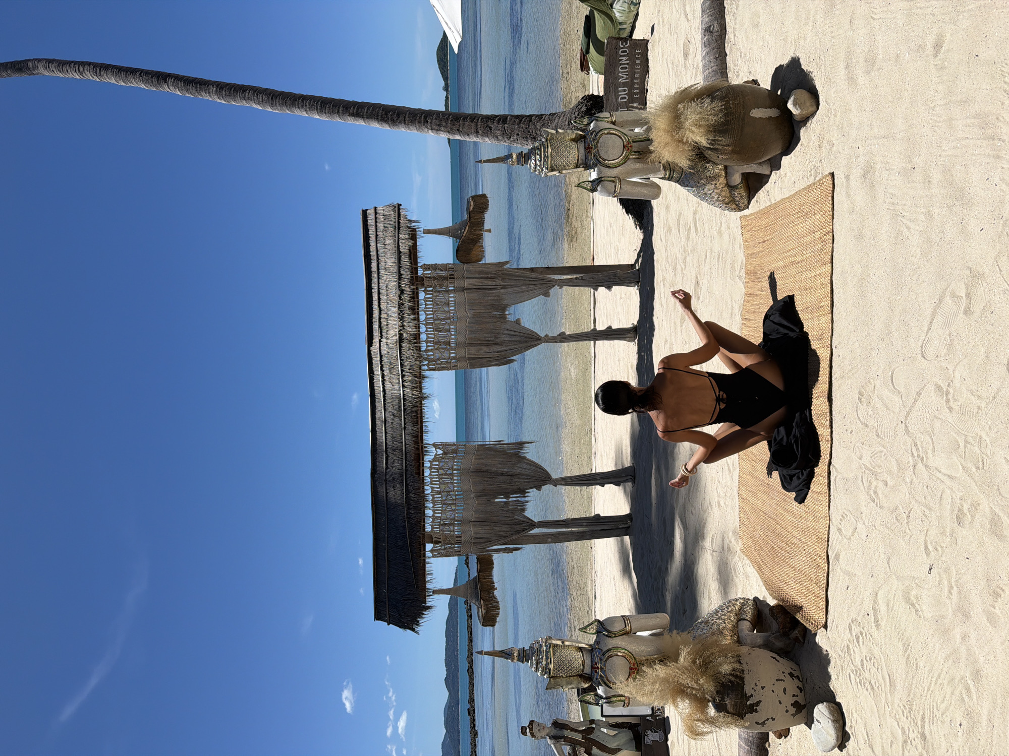 A woman in a black swimsuit is meditating on a woven mat on a sandy beach. The scene is set in Baan Thong Krut, Thailand. A tall palm tree trunk curves into the frame from the left, its rough texture contrasting with the smooth, clear blue sky. In the background, the shallow, calm ocean meets a distant, hazy shoreline.
To the right of the woman, a decorative structure with a thatched roof and woven drapes stands on the sand, casting a shadow. Nearby, an ornate, seated figure, possibly a statue or effigy adorned with intricate metallic details and a fluffy headdress, is partially visible. A sign with text is positioned behind this figure, but the text is mostly obscured.
The beach is composed of fine, light-colored sand with footprints visible. The sunlight suggests it is daytime, likely mid-morning or afternoon, given the clear sky and strong shadows. The overall atmosphere appears peaceful and serene, with the woman engaged in a moment of quiet contemplation by the sea. The presence of the statue and decorative elements hints at a cultural or resort setting.