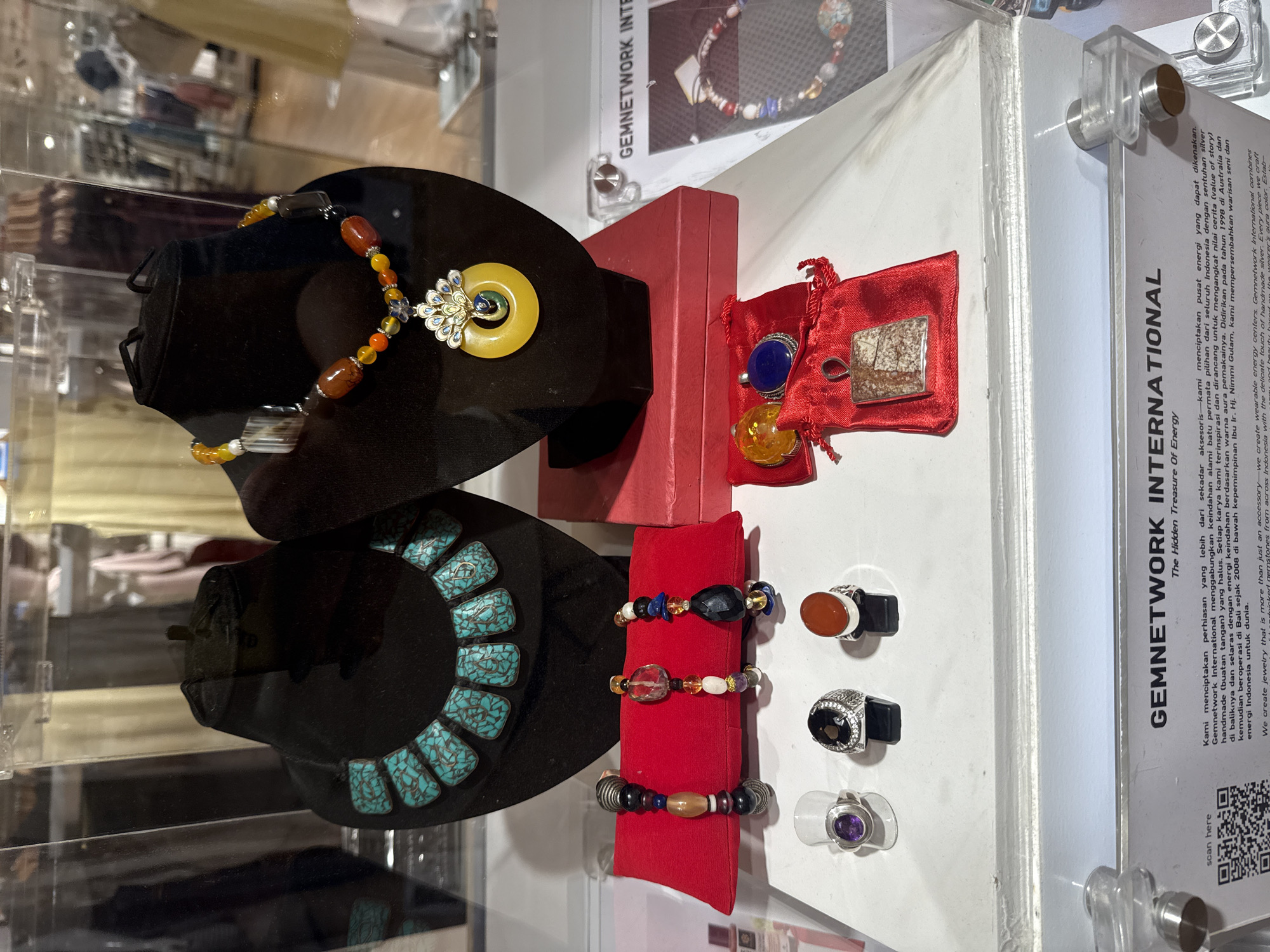 The image showcases jewelry displayed in a glass case, likely within the Gemnetwork International store, described on the informational sign. There are two black display busts, one adorned with a necklace featuring amber-colored beads and a yellow jade pendant, and the other with a turquoise necklace. A red box and a small red bag containing rings with gemstones are arranged nearby. Three individual rings are displayed on clear stands, and a red stand showcases three bracelets with various colored beads. A sign with the Gemnetwork International logo and text (including the phrase "The Hidden Treasure of Energy") is visible on the right side of the display. A QR code for the store is located at the bottom of the sign. The location is presumably within Denpasar, Indonesia.