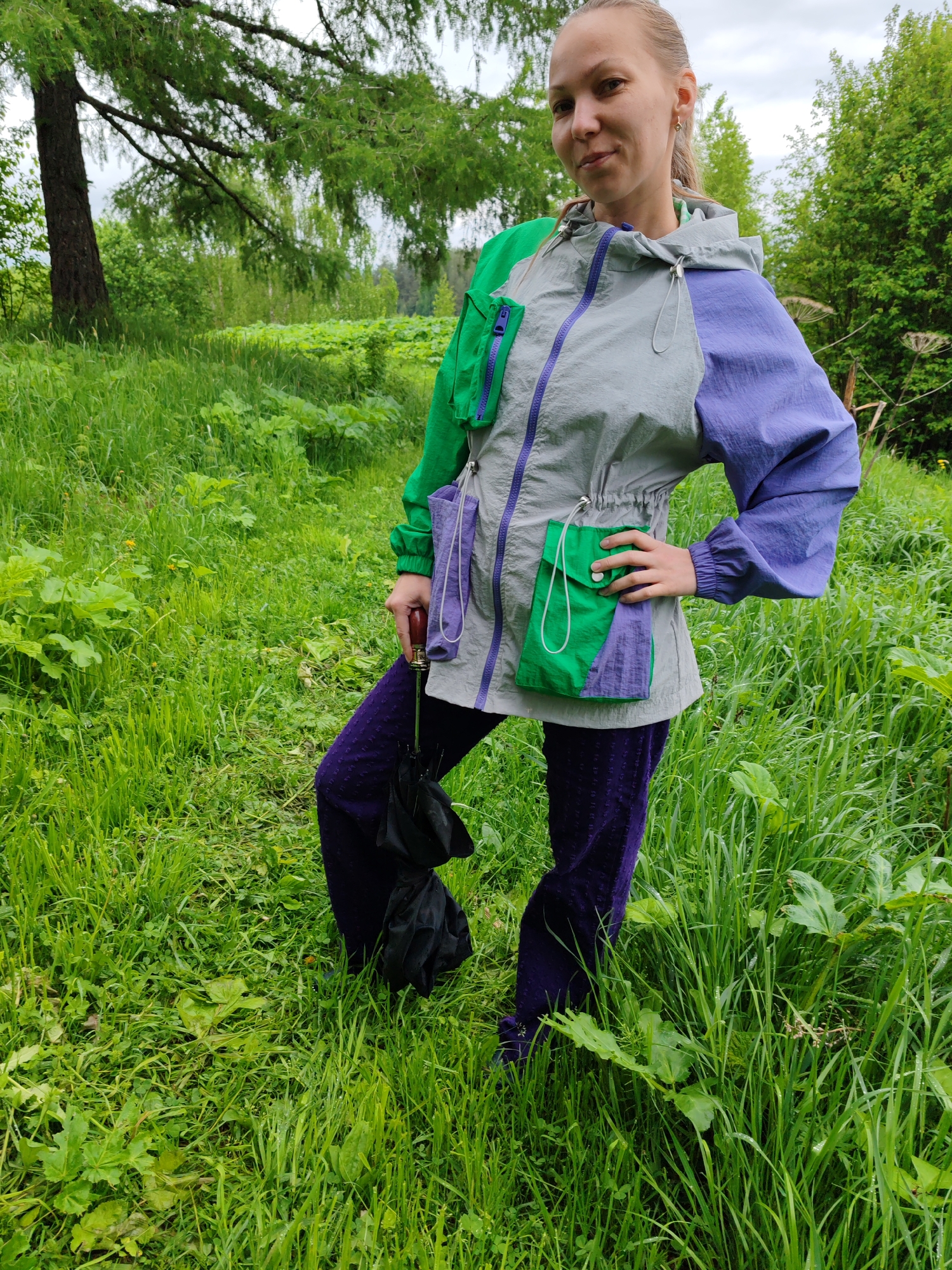The image depicts a woman standing in a grassy field in Чухлэм, Russia. She is fair-skinned with light-brown hair pulled back into a ponytail. She is wearing a multi-colored windbreaker jacket with green on the left shoulder, gray in the middle, and purple on the right shoulder, and purple pants. She has her left hand on her hip and her right leg raised. She is holding a black umbrella with a wooden handle. The background consists of green foliage, including trees and tall grass. The sky is overcast.