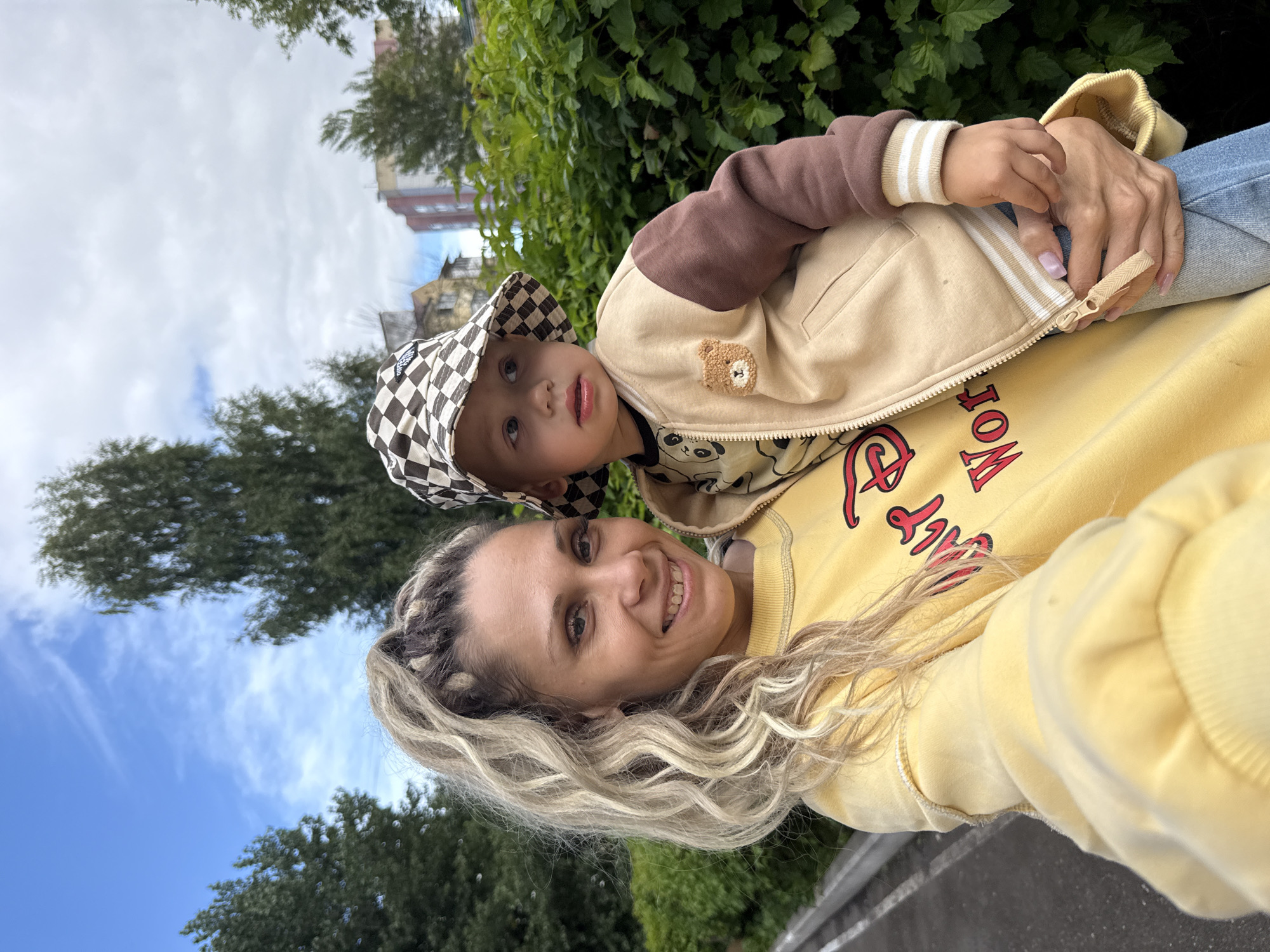In an outdoor urban setting in Rusanovo, Russia, a woman with blonde, wavy, high-ponytail hair and a yellow sweatshirt is holding a young child. The child is wearing a beige and brown jacket with a checkered hat. They are positioned in front of a dense green hedge with trees and buildings visible in the background under a partly cloudy sky. The woman is smiling at the camera, and the child is looking to their right. The woman's left arm is around the child, and her right hand is visible holding the child. The image appears to be a selfie.